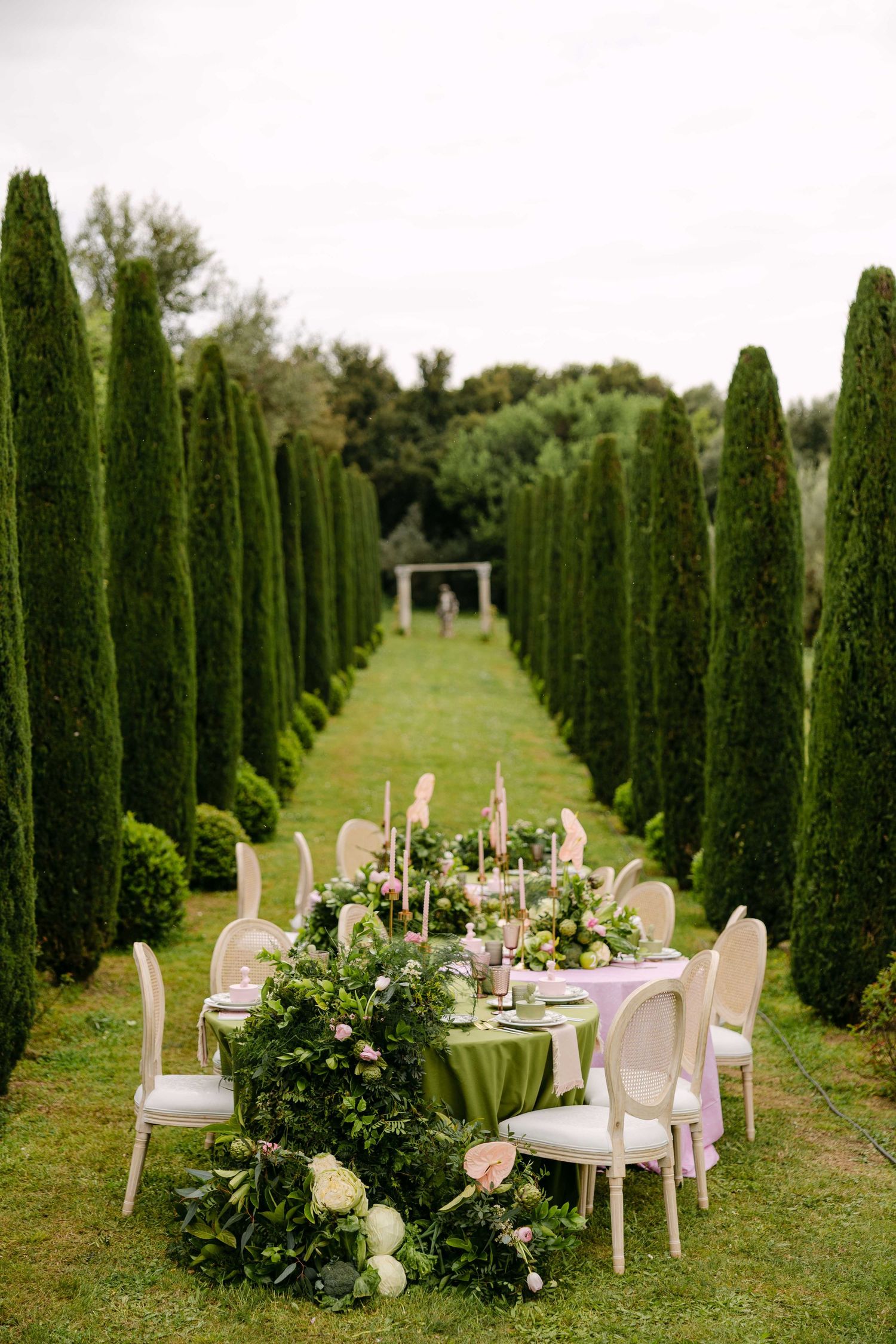Elegant garden dinner table with green tablecloth and floral arrangements set between manicured cypress trees in formal garden.