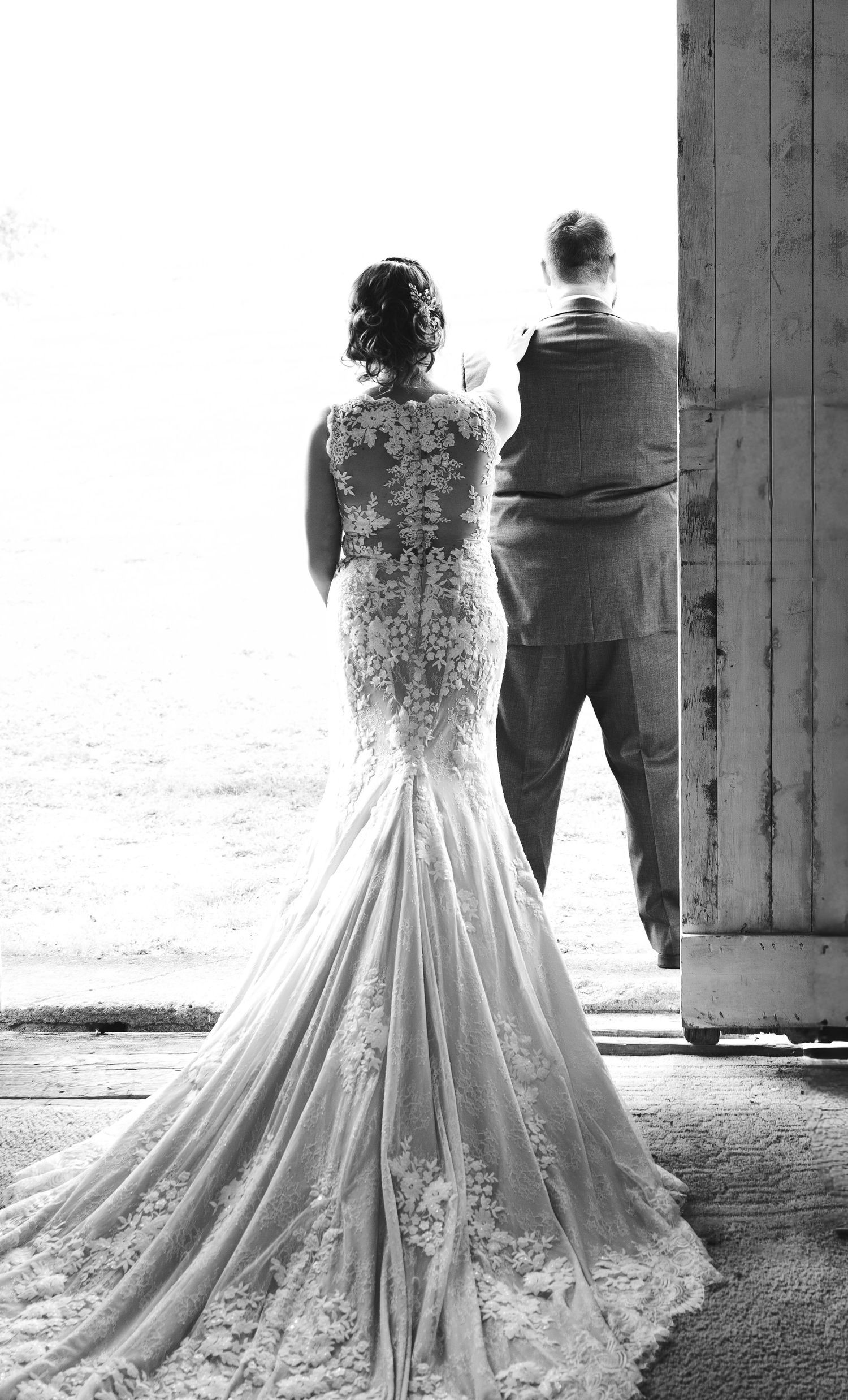 Black and white rear view of bride in lace mermaid gown with long train beside groom in barn.