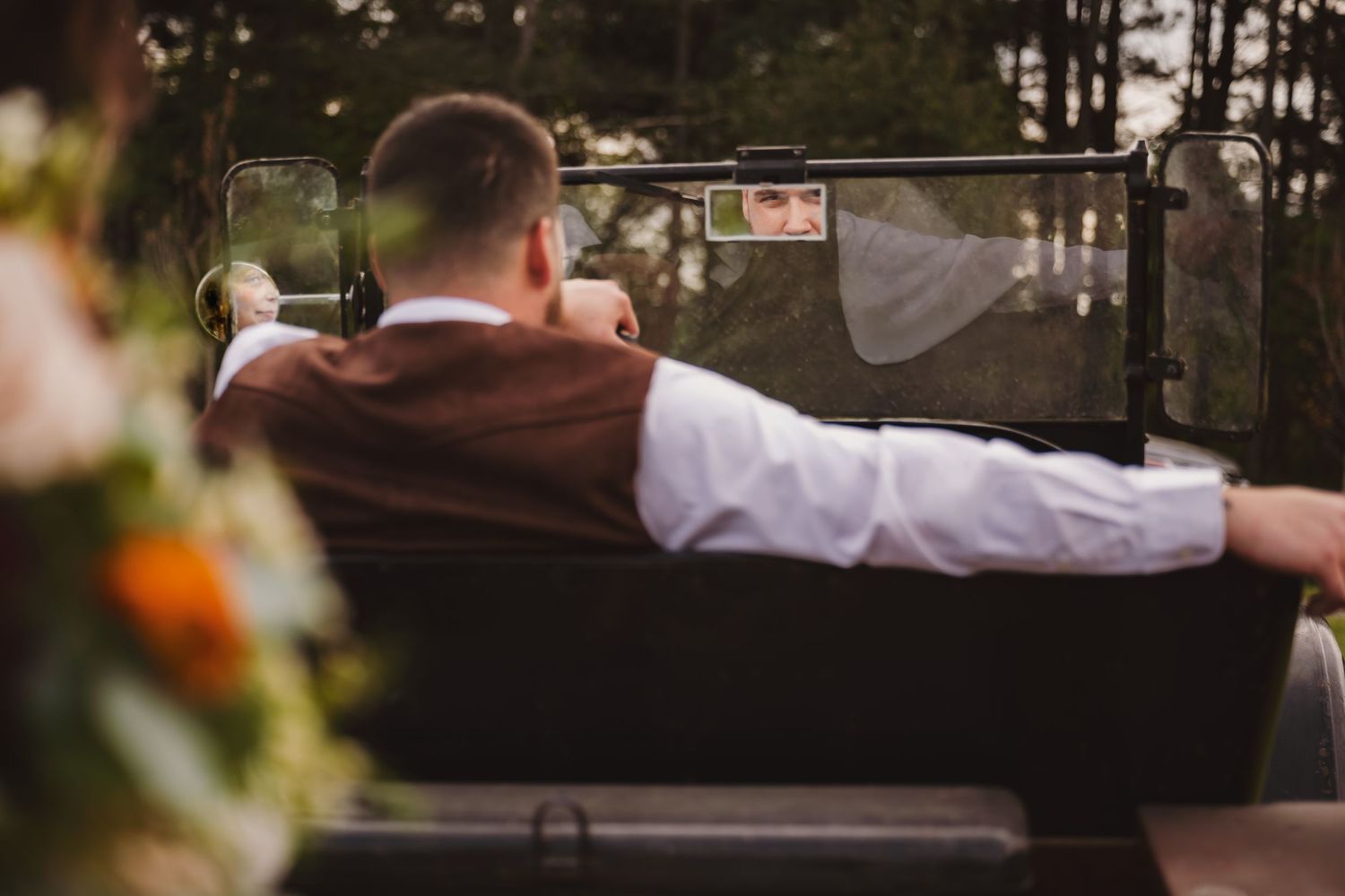 Groom in brown vest sits in vintage car as bride's reflection appears in rearview mirror.