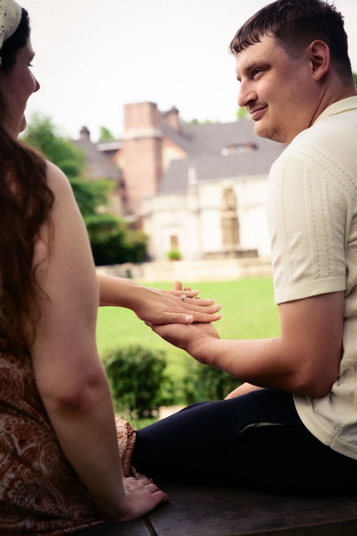 A couple sits outdoors holding hands near a historic building, appearing to share a romantic engagement moment.