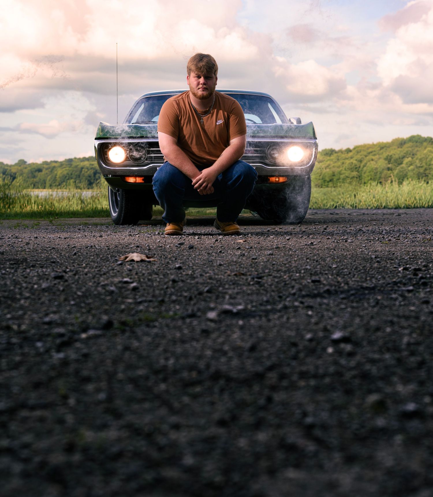 Man crouching in front of a classic black muscle car on a rural road at dusk with dramatic cloudy sky.