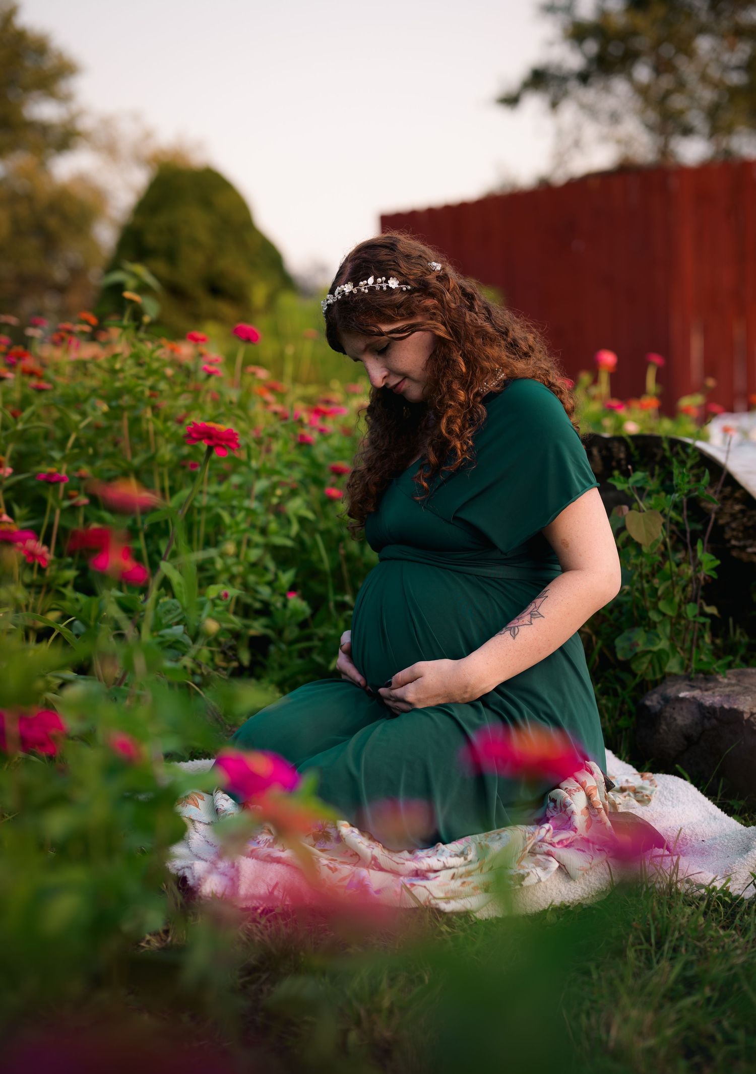 Pregnant woman in green dress sitting among pink flowers wearing floral crown during golden hour maternity photoshoot.