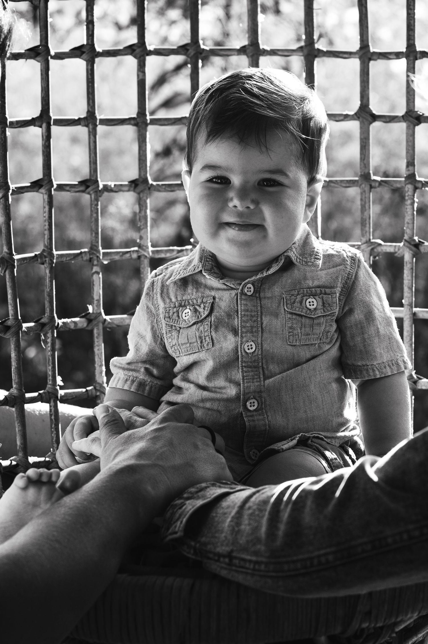 Black and white portrait of a smiling toddler boy in a denim shirt sitting on an adult's lap near a metal fence.