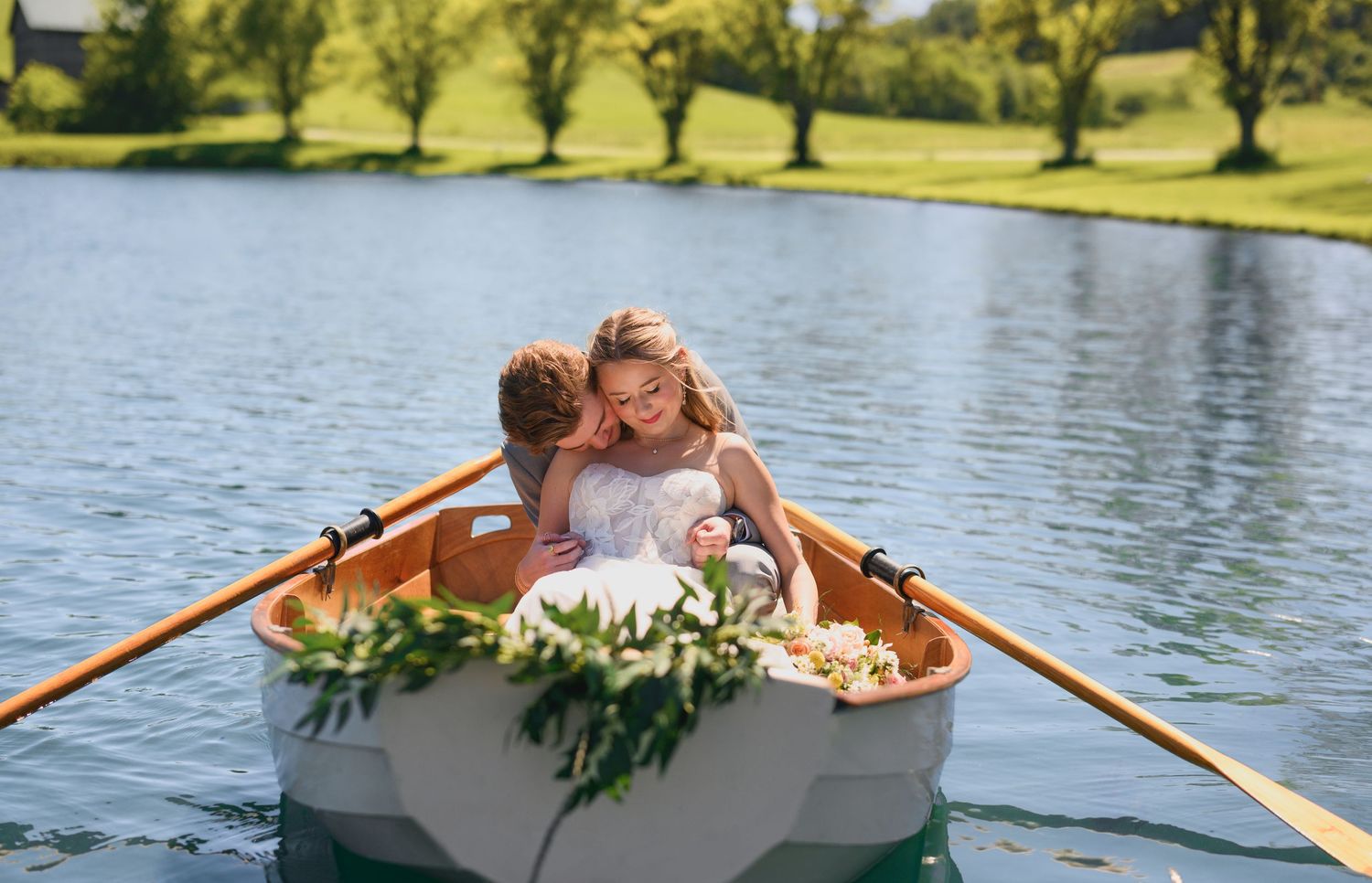 Couple embracing in a flower-adorned rowboat on a serene lake, surrounded by lush greenery on a sunny day.
