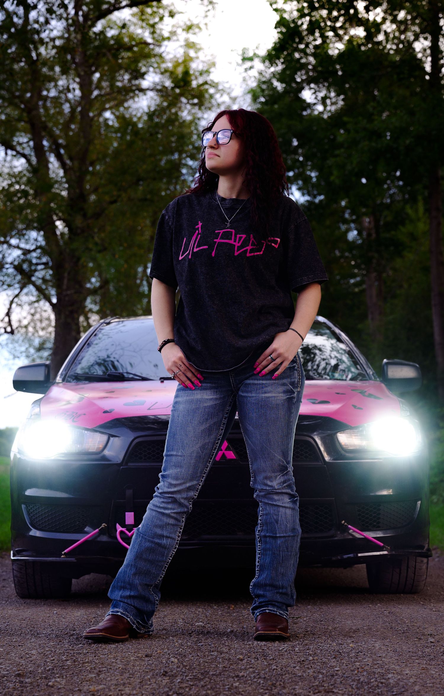 Young woman in black Lil Peep tee and jeans poses confidently in front of a black and pink sports car at dusk.