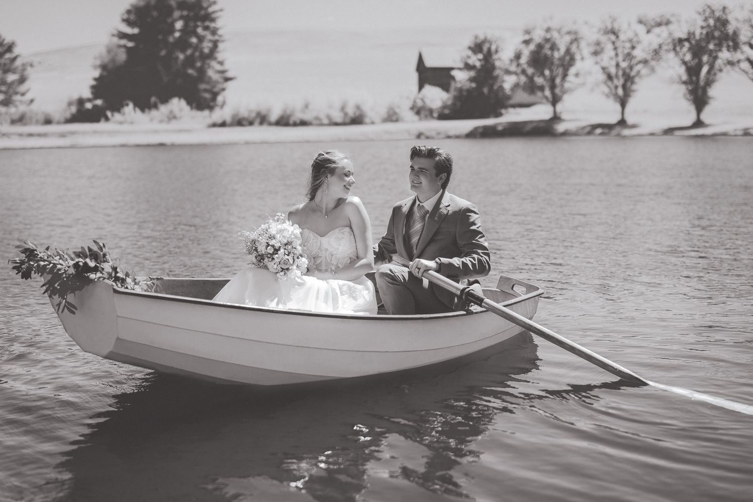 Black and white photo of a bride and groom rowing a small boat on a calm lake on their wedding day.