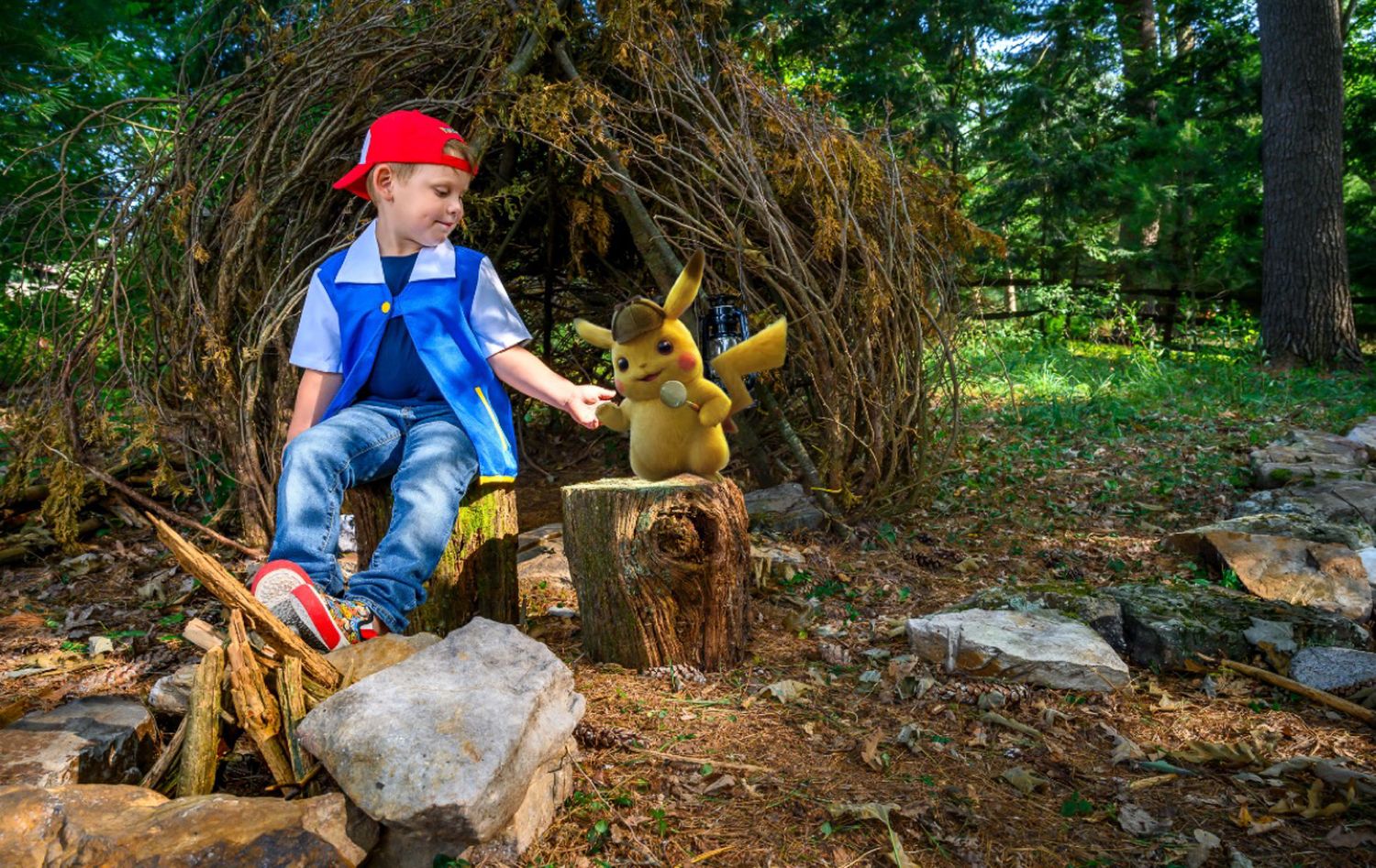 Young boy dressed as Ash Ketchum poses with a Pikachu statue in a woodland fairy tale setting.