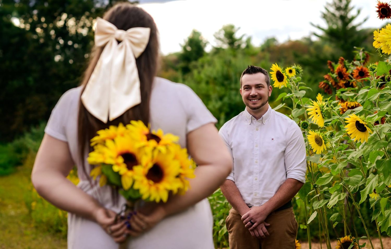 Couple in sunflower field, woman holding yellow sunflower bouquet with bow, man smiling in white shirt.