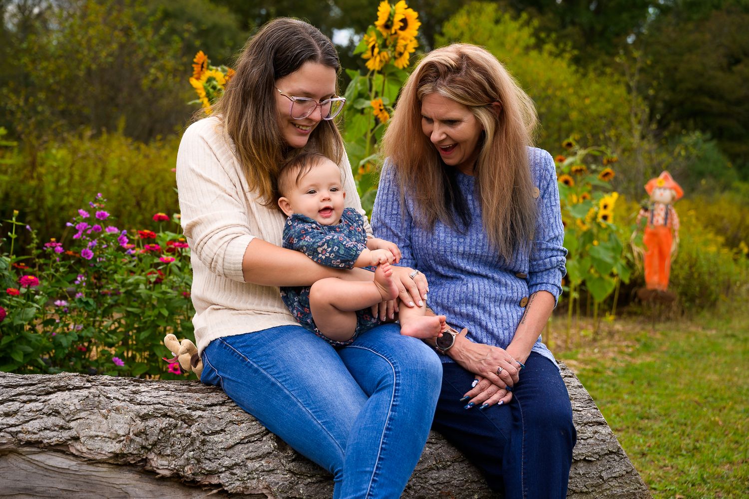 Two women and a baby sit on a rock surrounded by sunflowers and colorful fall garden blooms outdoors.