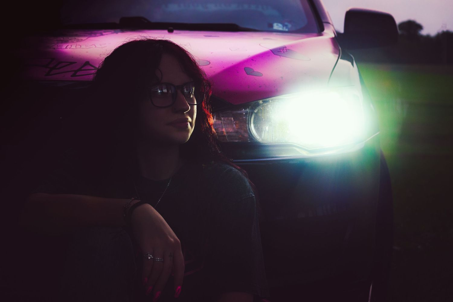Woman with glasses sitting beside a pink car illuminated by bright headlights at dusk.