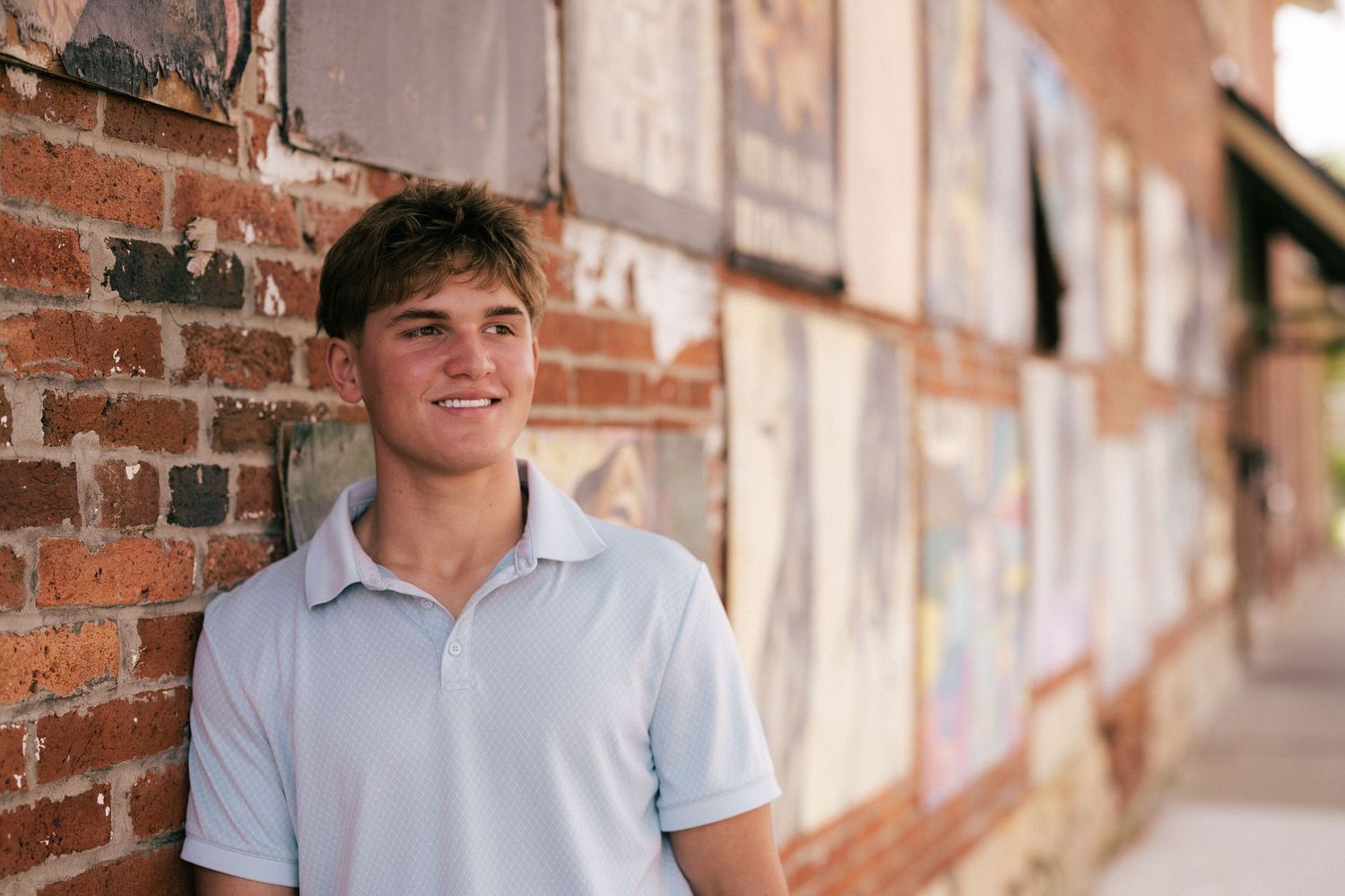 Smiling teenage boy in white polo shirt leaning against a brick wall outdoors in a casual portrait.