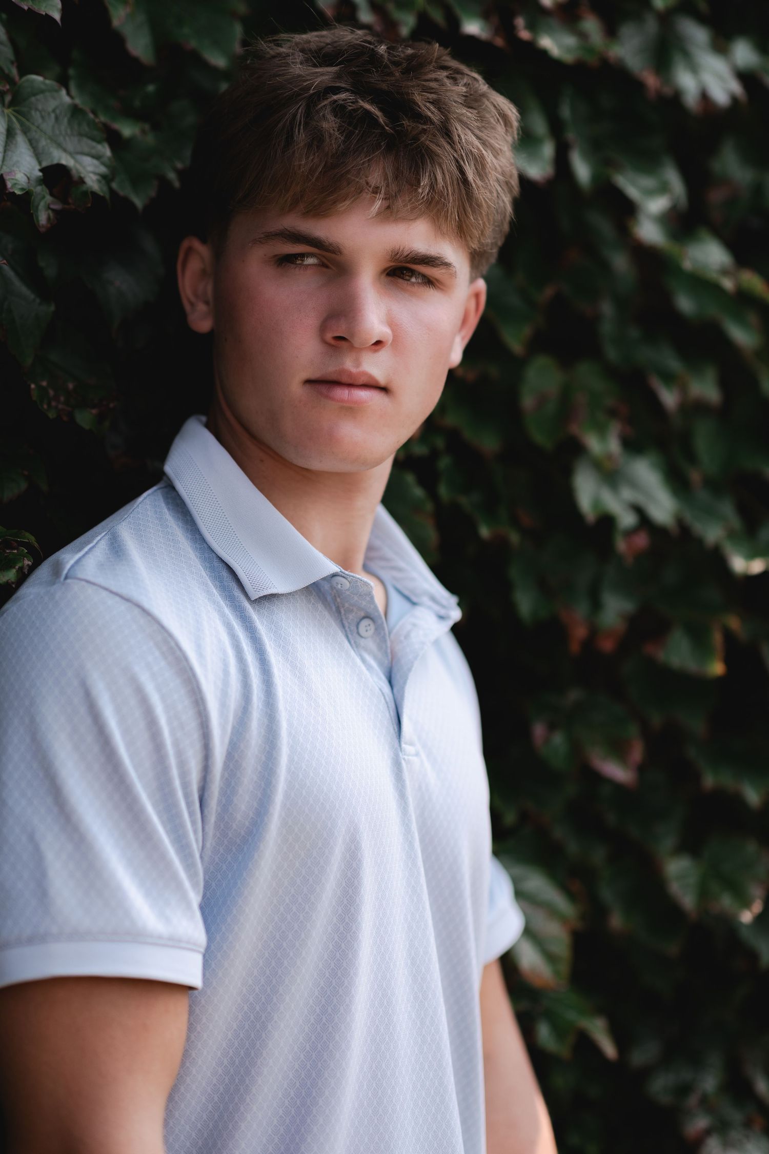 Young man in white polo shirt posing outdoors against lush green ivy wall in natural light portrait.