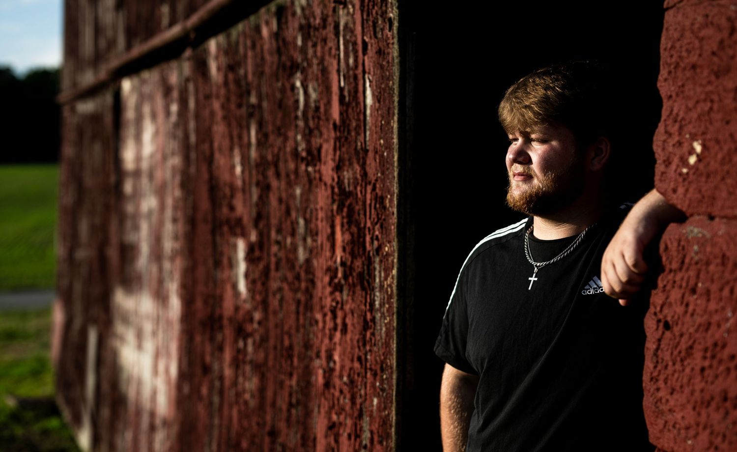 Young man in black t-shirt and cross necklace leaning against a rustic red barn in sunlight.