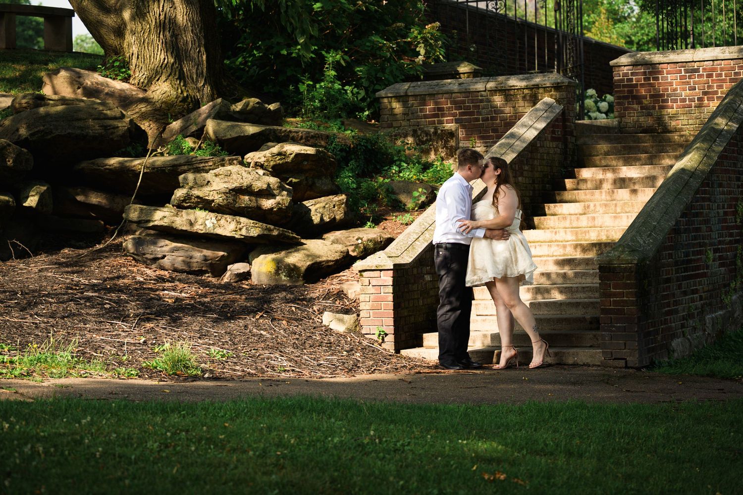 Couple embracing romantically near brick steps and stone wall in a lush green park setting.