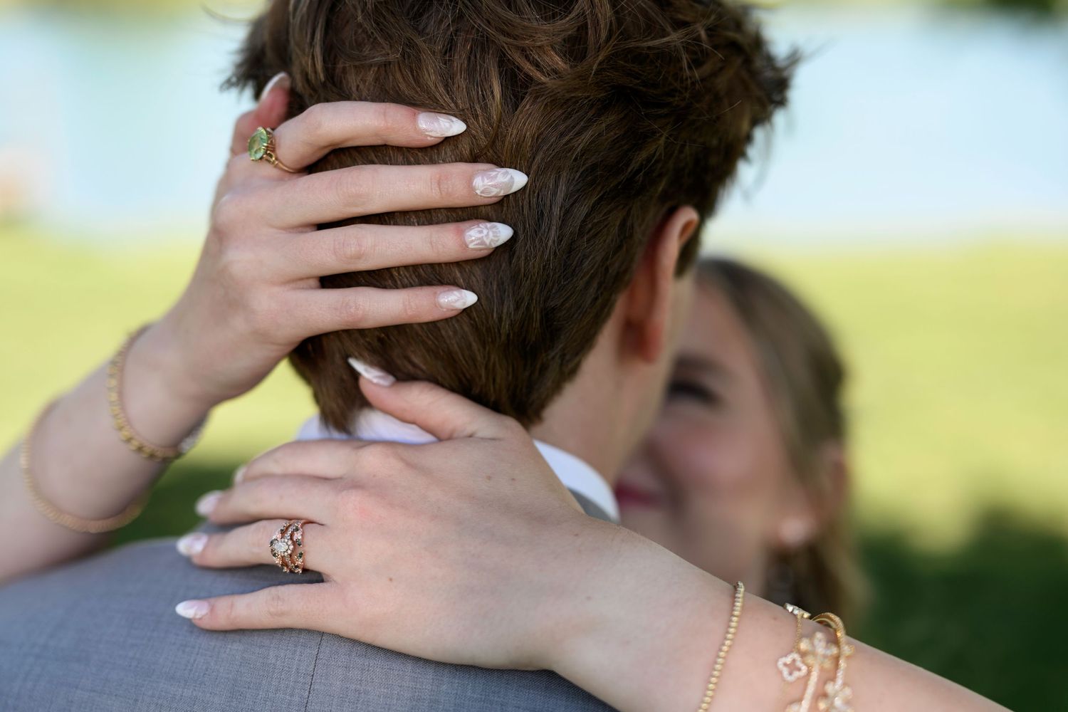 Bride with long nails and gold jewelry embracing groom in gray suit outdoors at wedding ceremony.