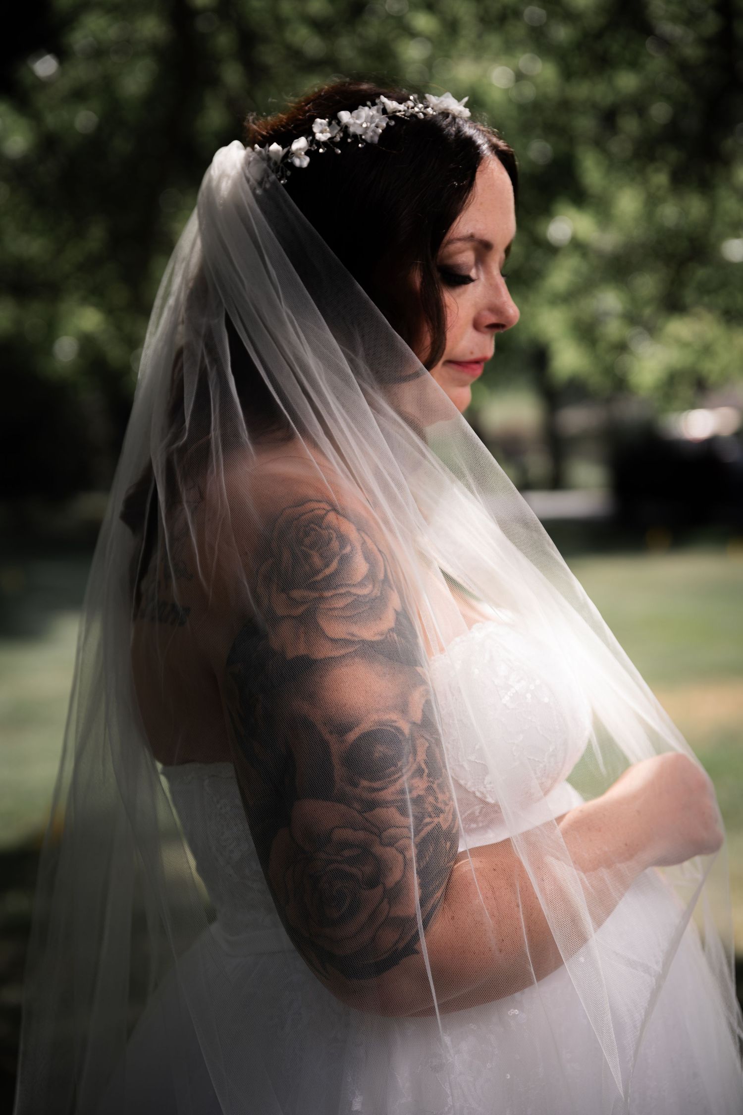 Tattooed bride with rose sleeve tattoo wearing veil and floral crown in soft outdoor lighting.