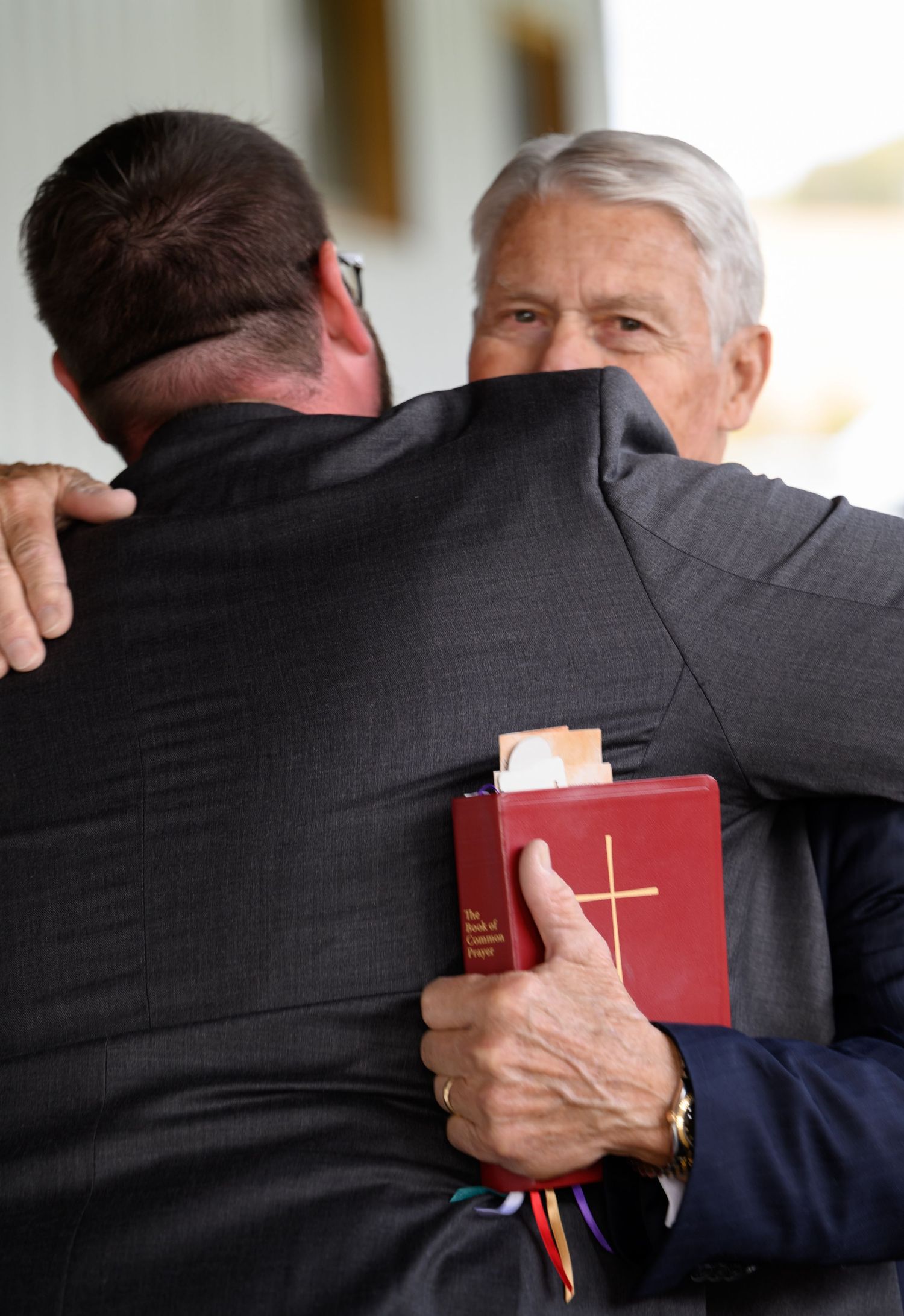 Officiant holding red Bible embraces groom in heartfelt hug after outdoor wedding ceremony.