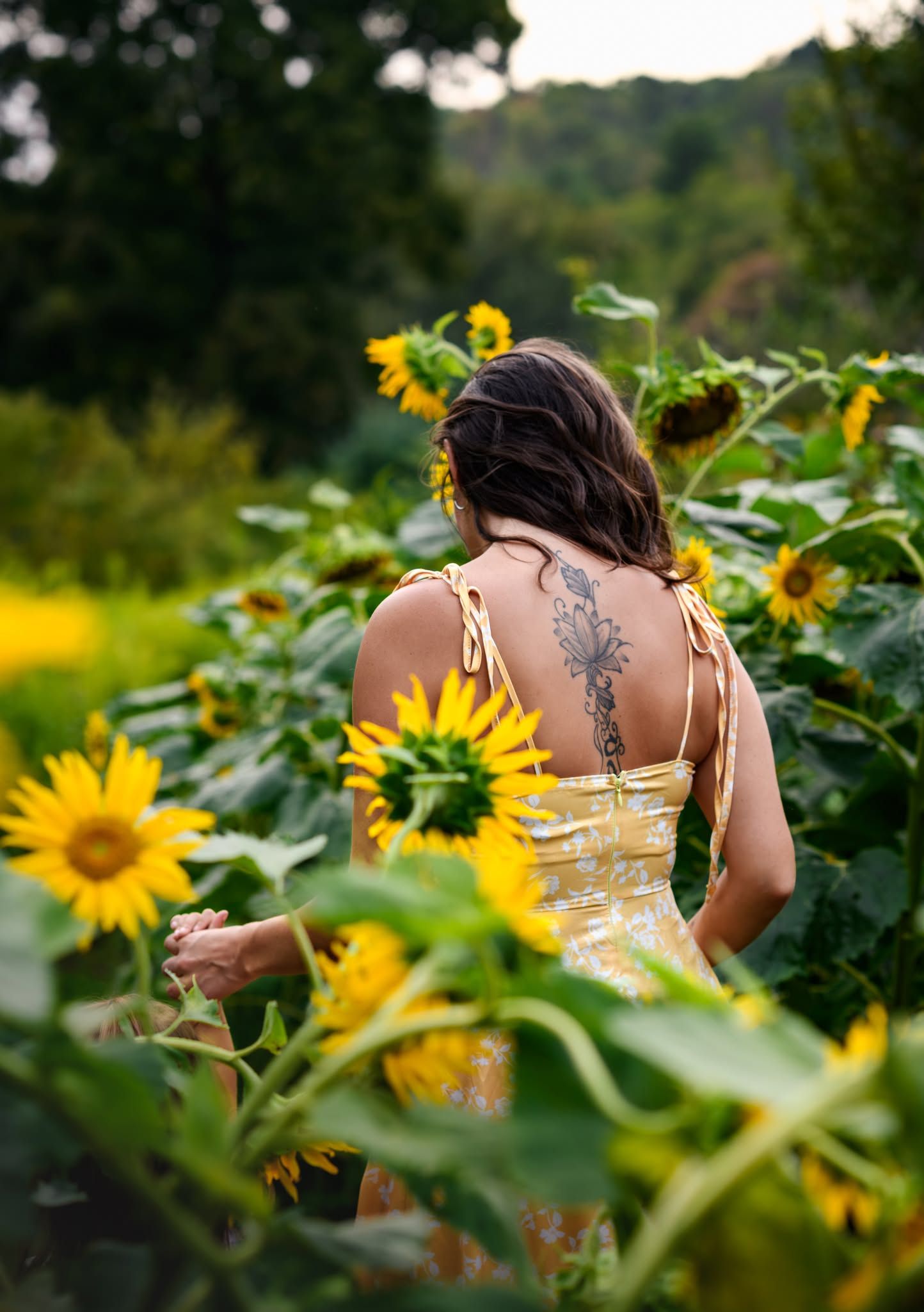 Woman with back tattoo in yellow sundress walking through vibrant sunflower field surrounded by lush green foliage.