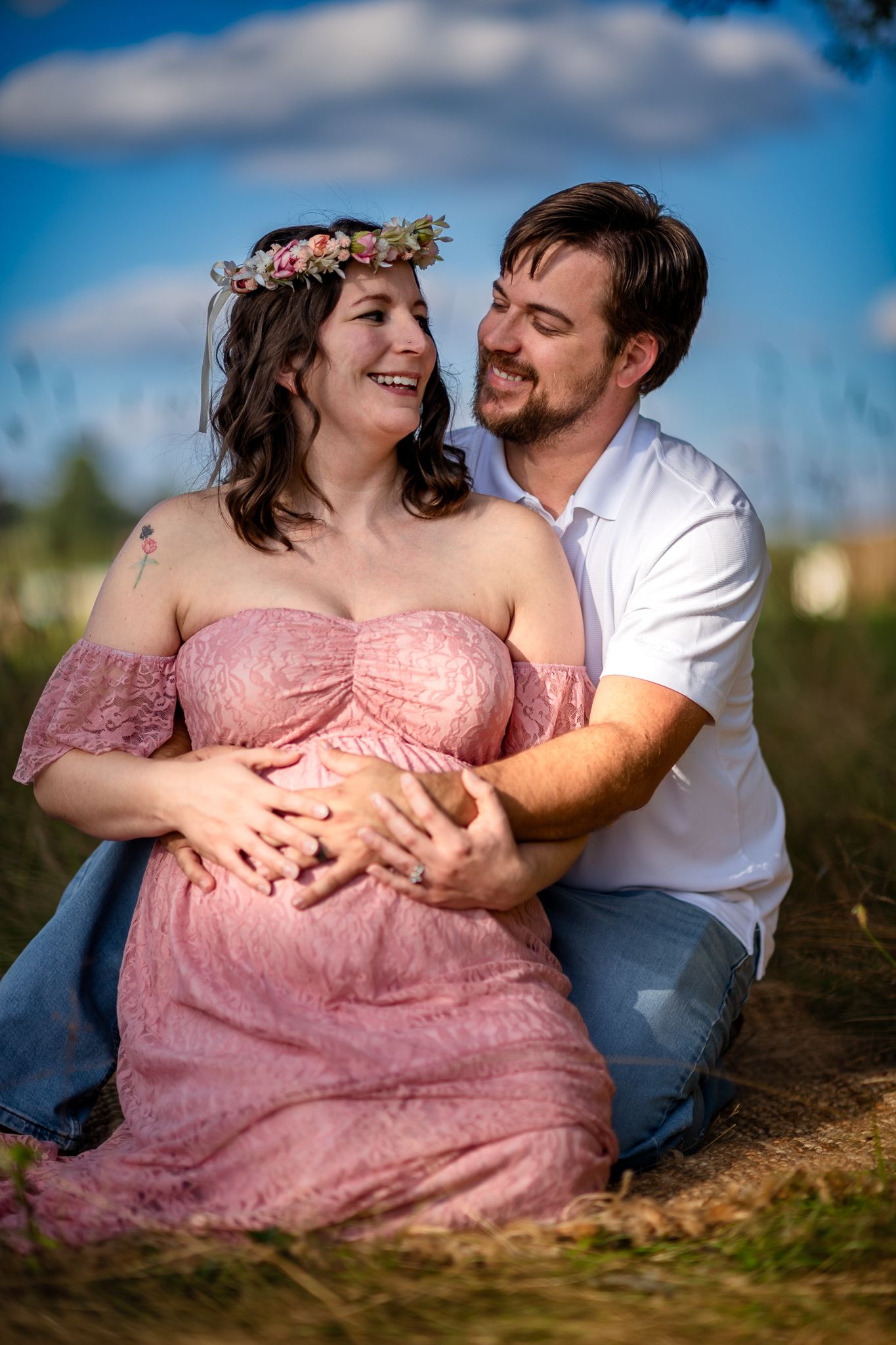 Couple sharing a joyful maternity photo shoot outdoors, woman in pink lace gown and floral crown, man embracing her baby bump.