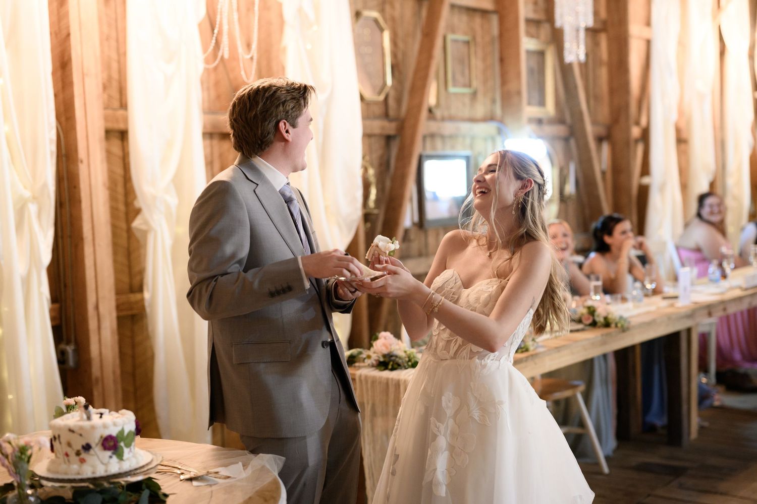 Bride and groom laughing together during wedding cake cutting ceremony inside a rustic barn reception venue.