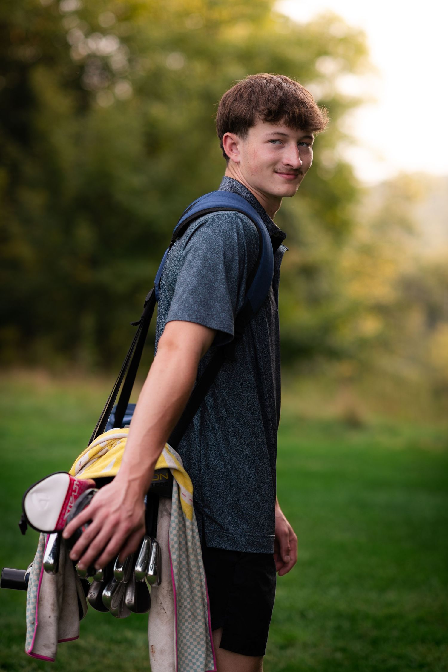 A teenage boy smiles while carrying a golf bag with clubs on a lush green golf course.