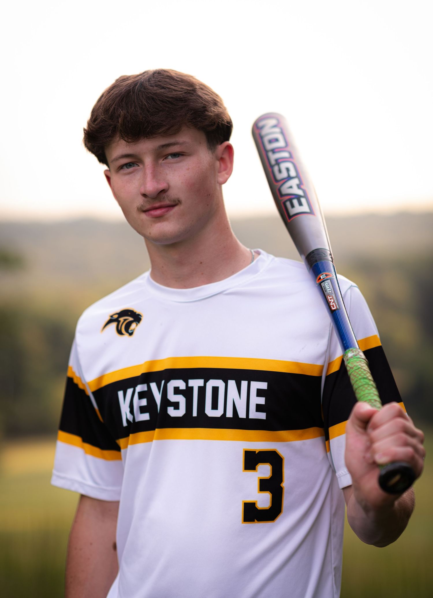 Keystone number 3 baseball player holding an Easton bat, wearing black and gold uniform outdoors.