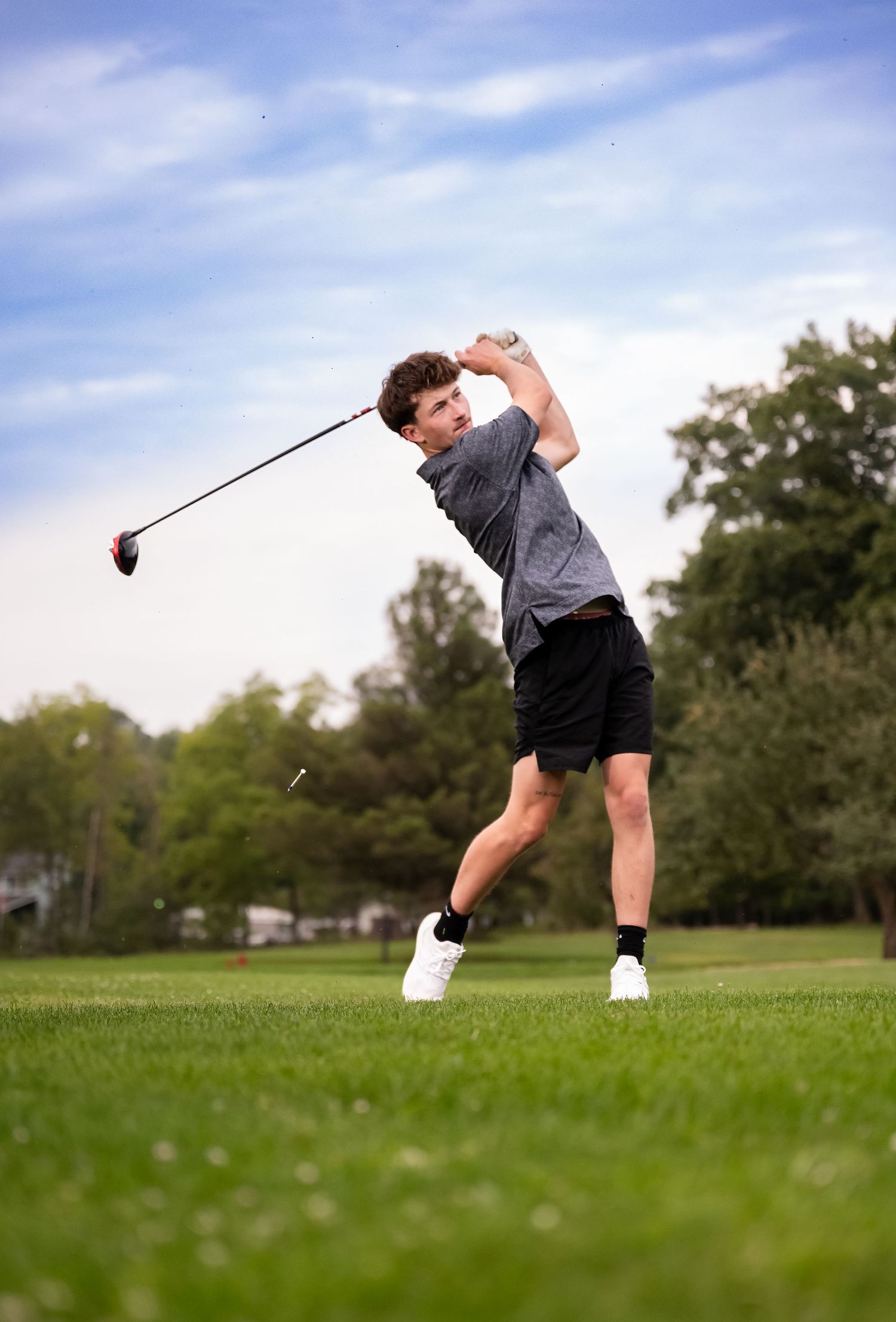 Young golfer follows through on a powerful drive swing on a lush green golf course under a partly cloudy sky.
