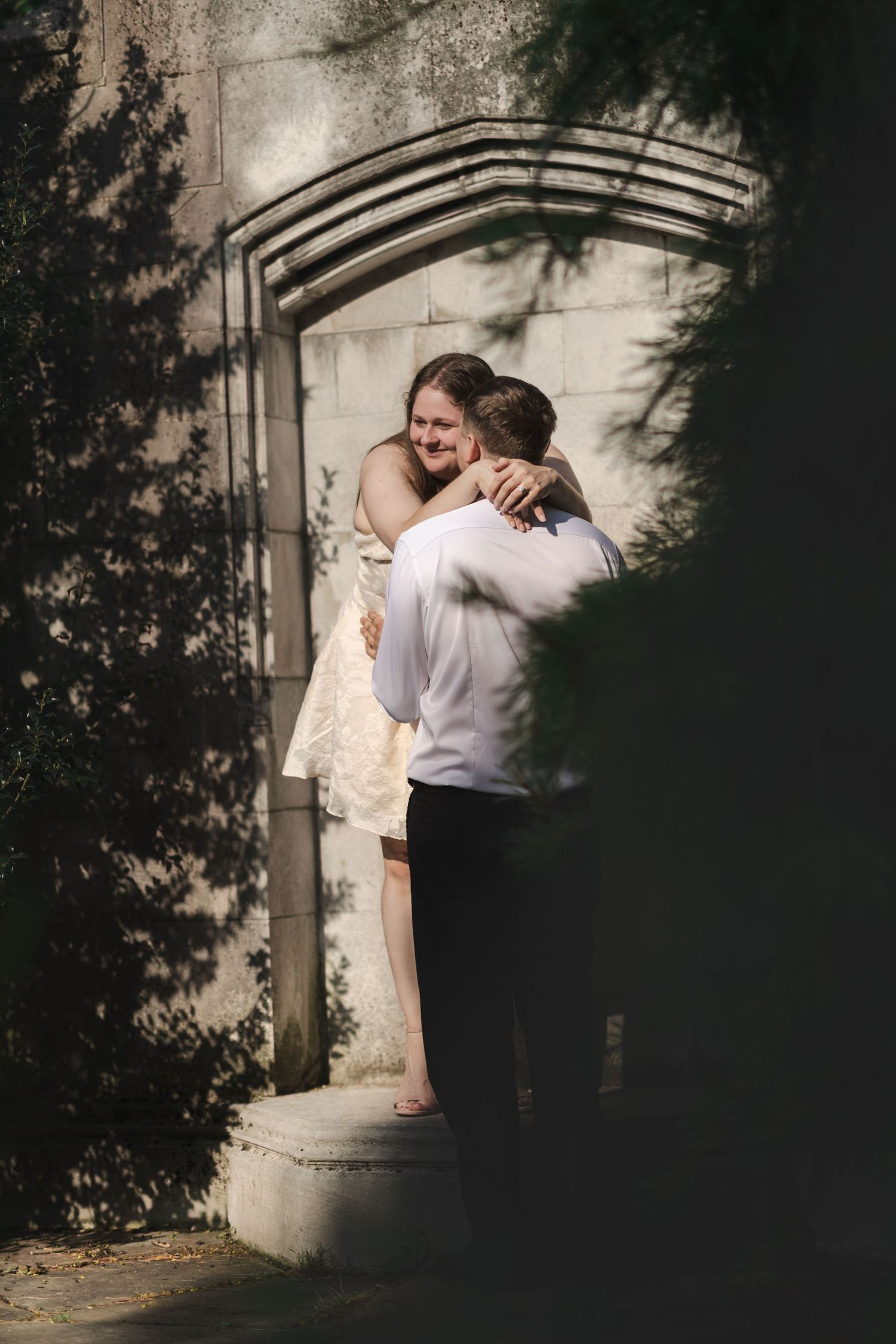 A groom lifts his smiling bride in a warm embrace on stone steps, framed by lush greenery and an arched wall.
