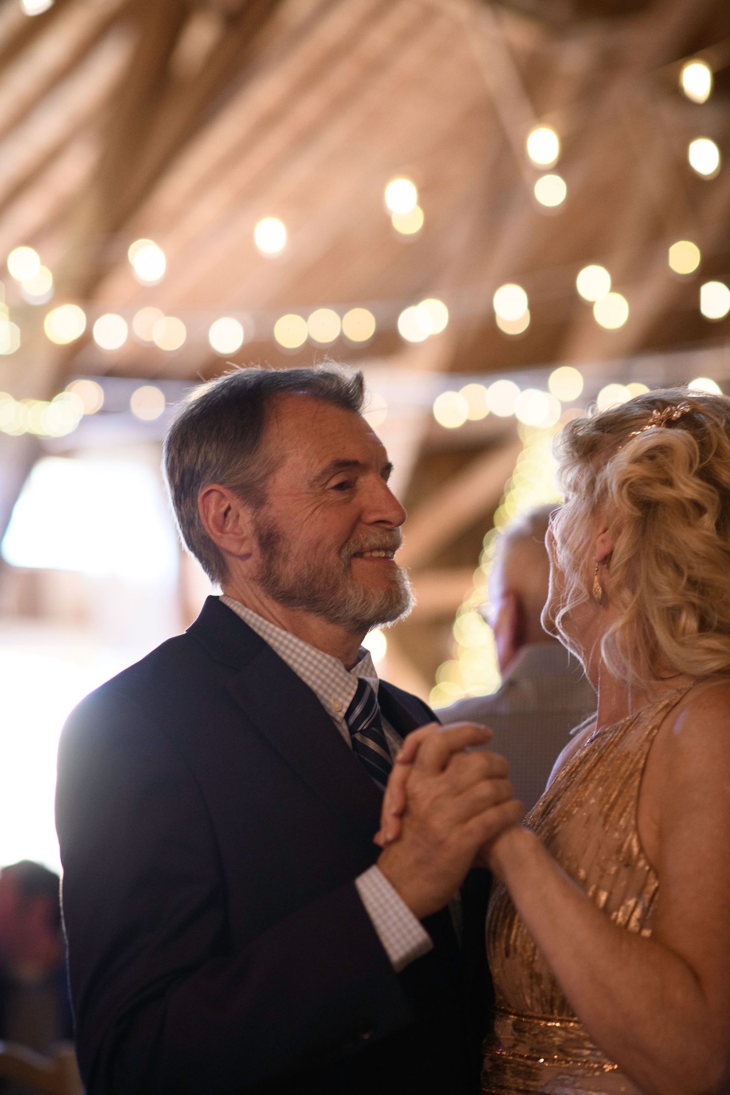 Mature couple sharing a romantic dance at a wedding reception under warm fairy lights in a rustic barn venue.