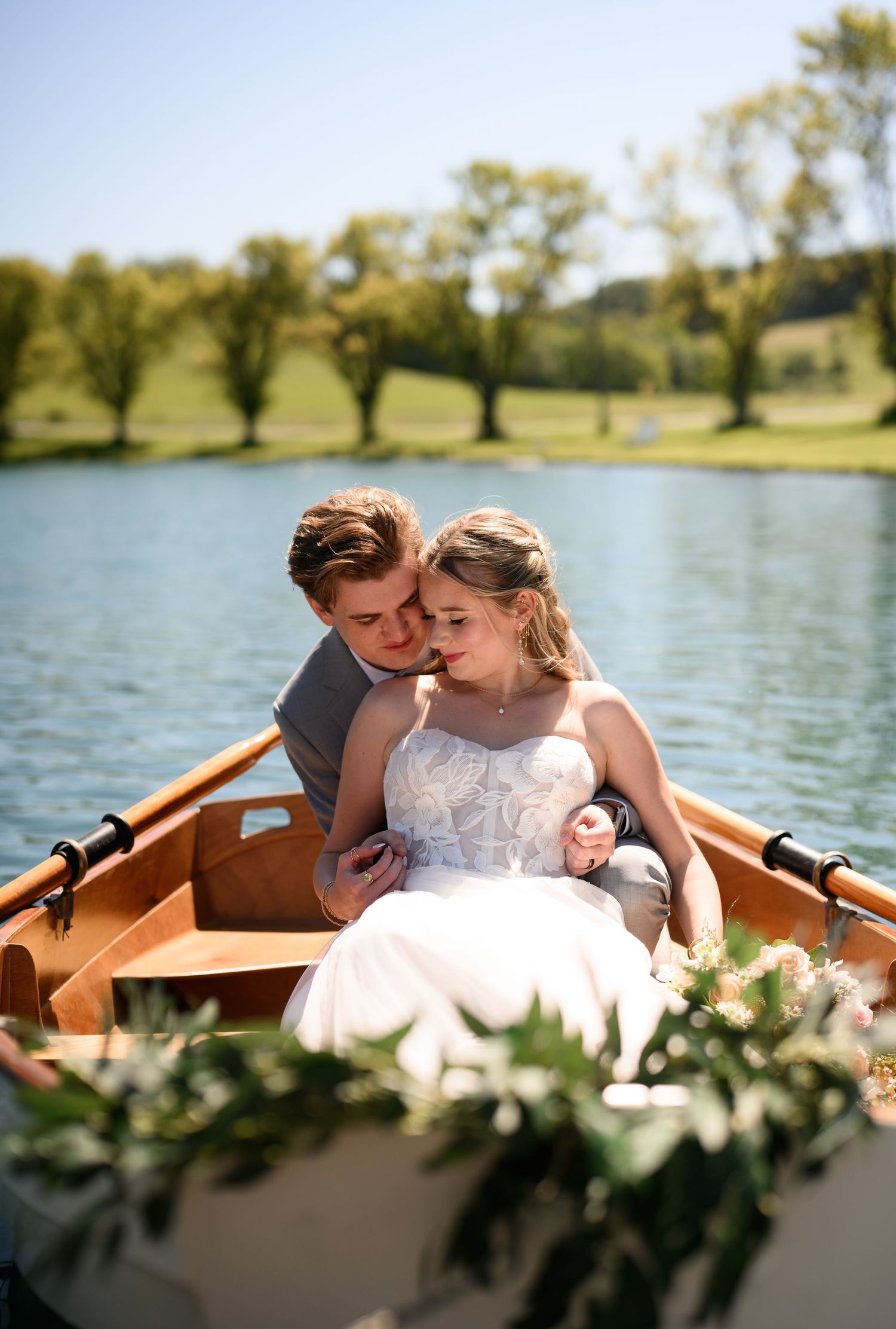 Bride and groom sharing a romantic moment in a wooden rowboat on a serene lake, surrounded by lush greenery.