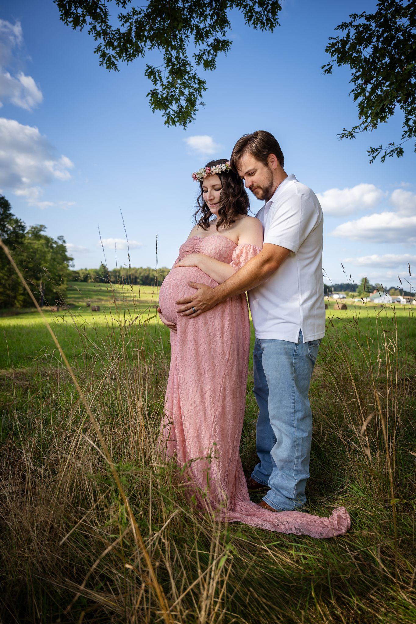 Expecting couple standing in green field, woman in pink maternity gown with floral crown, man in white shirt.