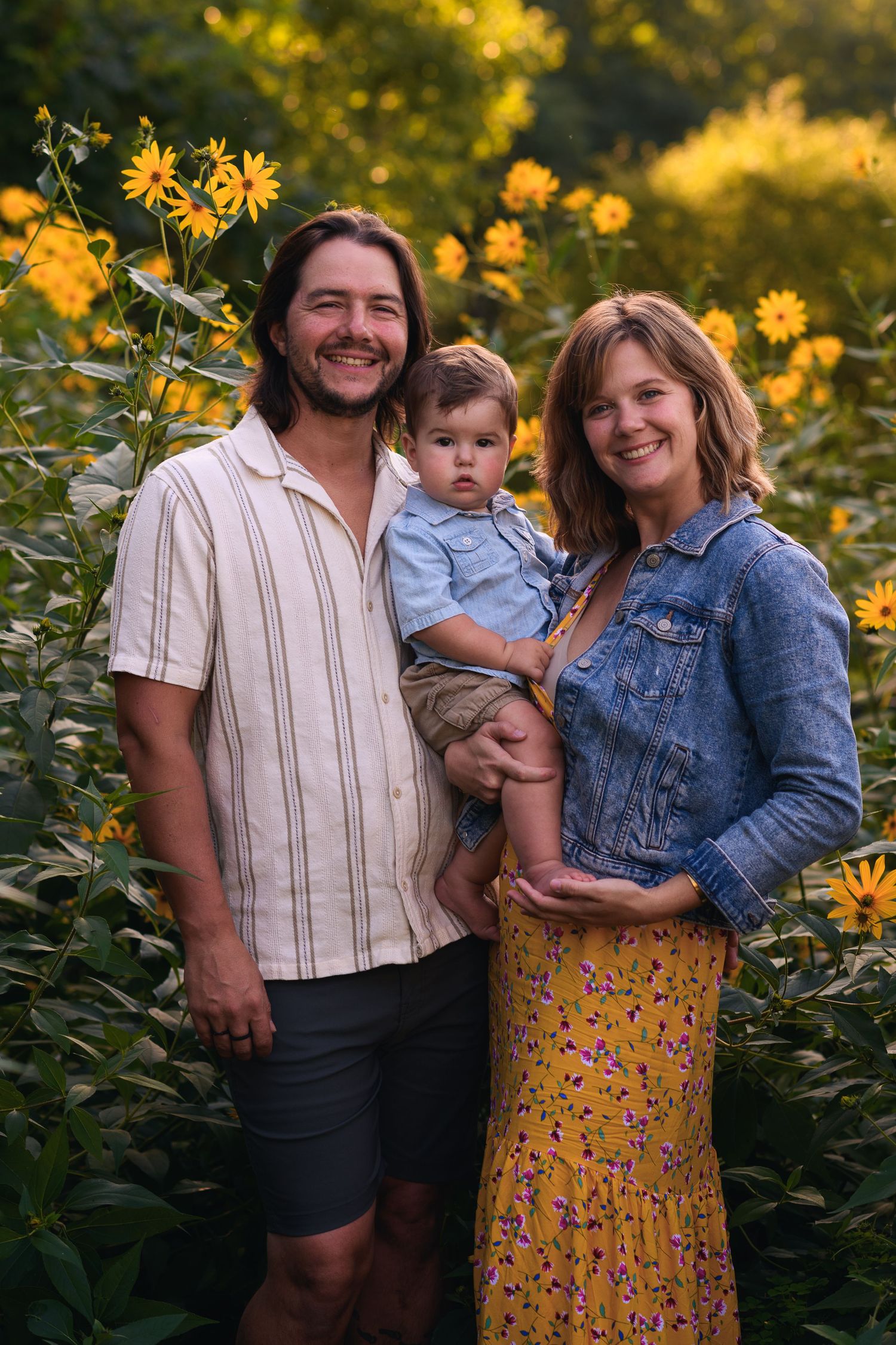 Happy family with baby posing in a sunny yellow wildflower field, parents smiling outdoors in casual summer attire.