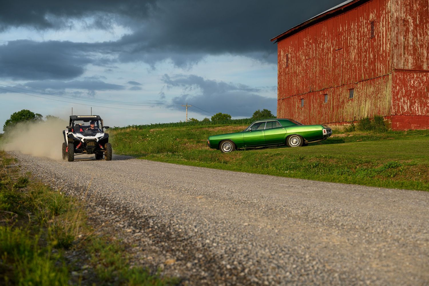 A green classic muscle car races a white RZR UTV on a gravel road beside a red barn under stormy skies.
