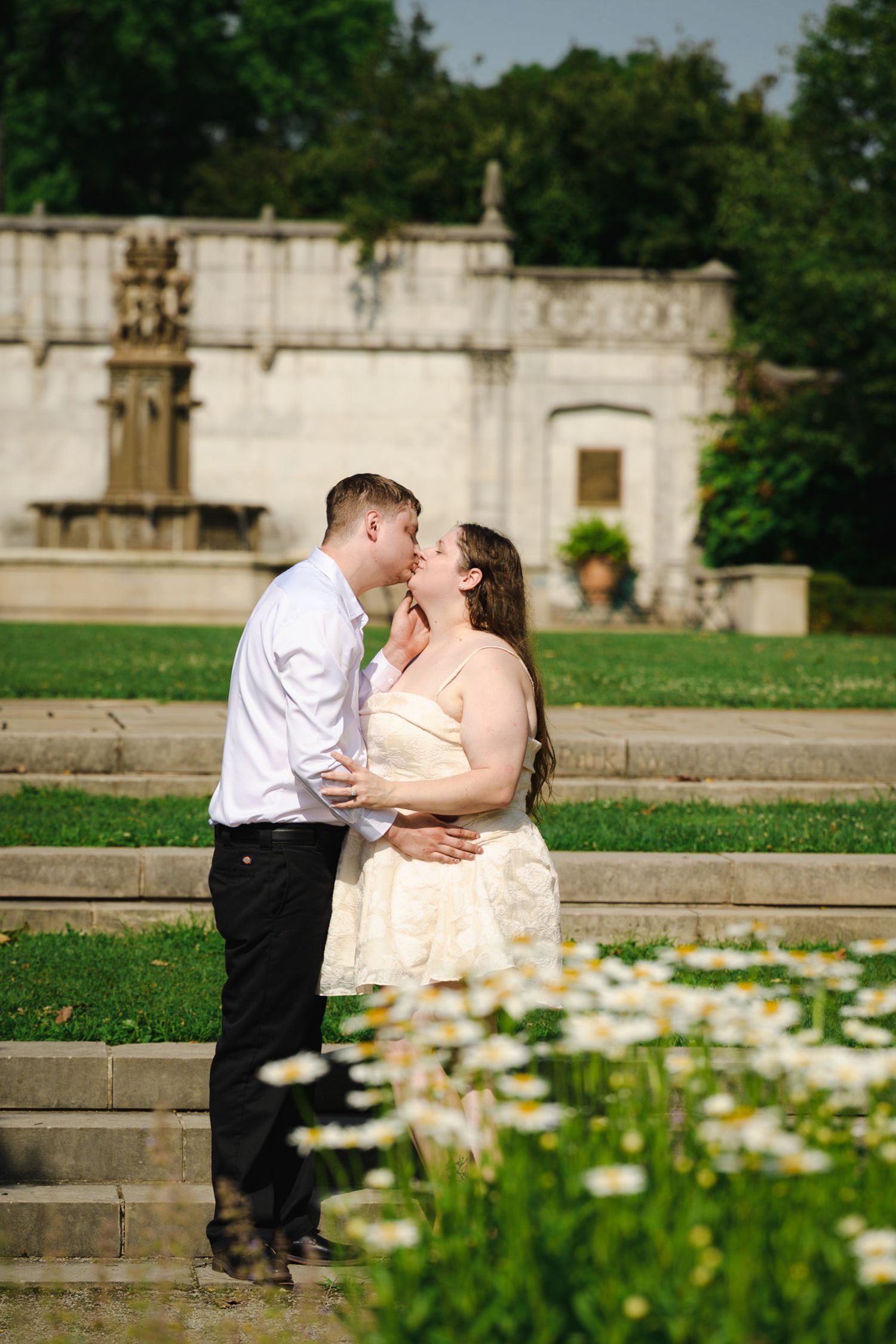Couple sharing a romantic kiss in a sunlit garden with white daisy flowers and a stone monument backdrop.