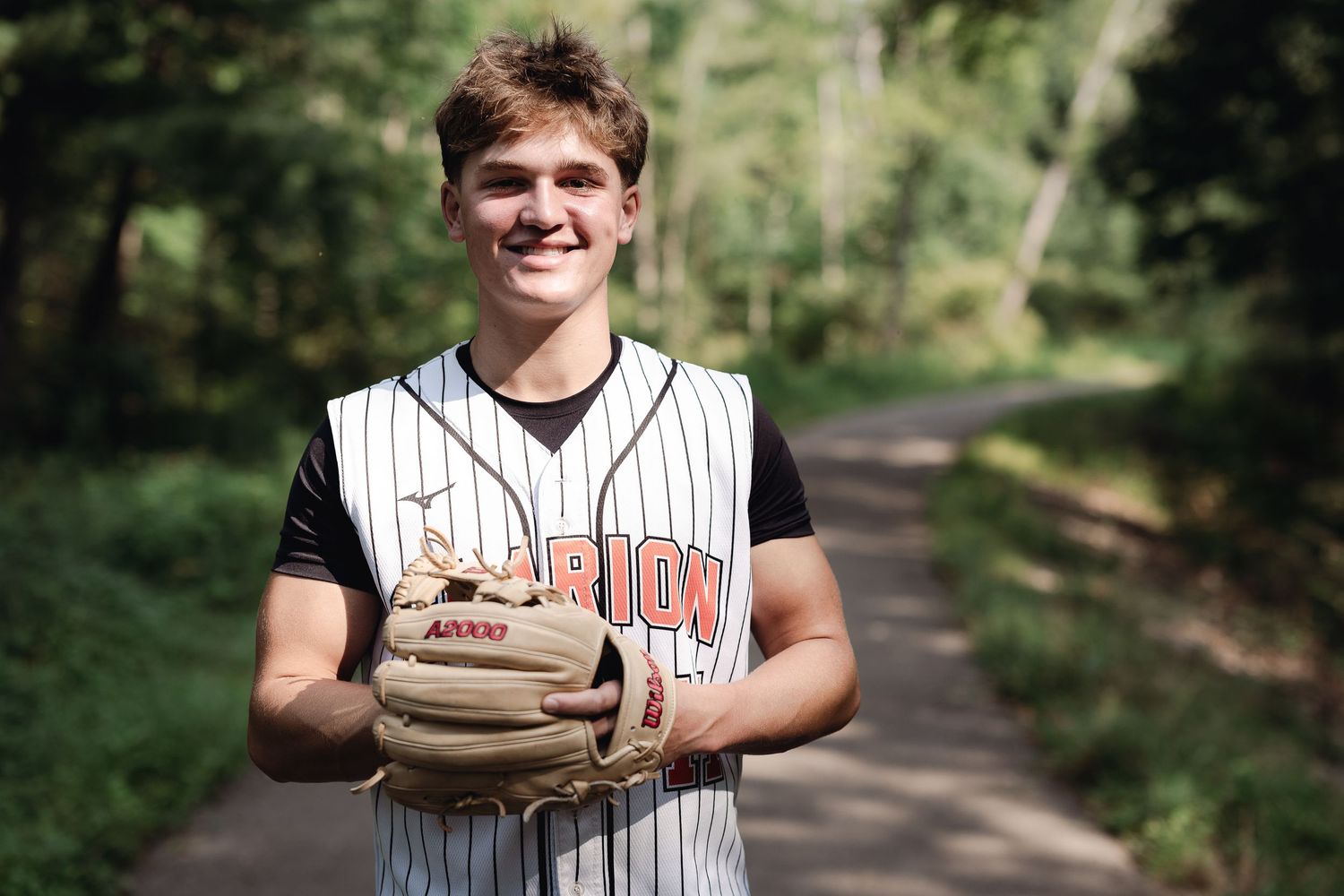 Young baseball player in Marion jersey smiling on a wooded path, holding a fielding glove.
