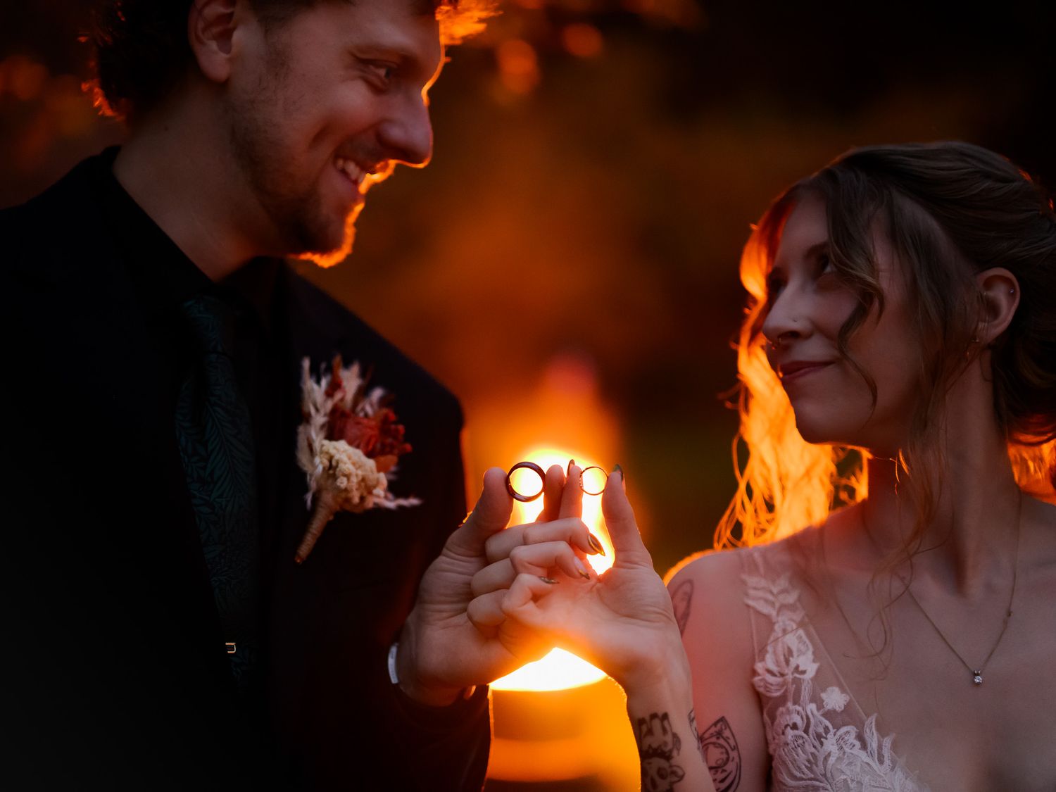 Couple sharing wedding rings by candlelight at dusk, groom in black suit, bride in lace gown with tattoos.
