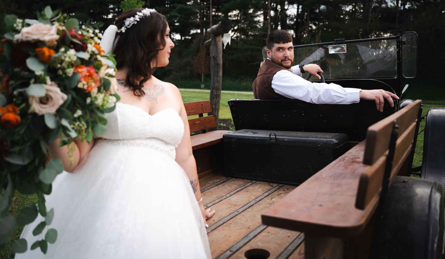 Bride holding bouquet stands on vintage wooden wagon as groom relaxes during rustic wedding.