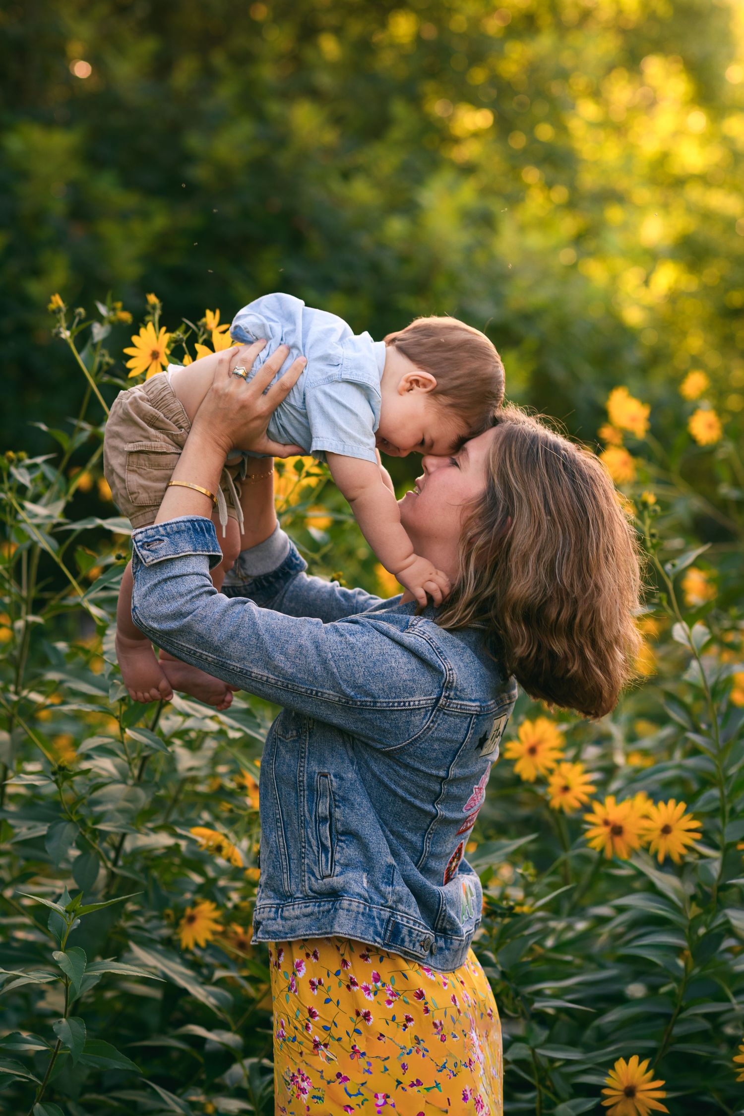Mother lifting baby in sunflower field, wearing denim jacket and yellow floral skirt, sharing a tender nose-to-nose moment.