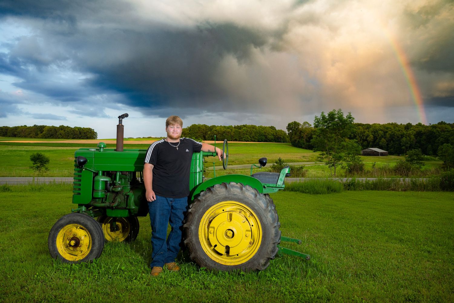 A man stands proudly beside a green John Deere tractor on a farm with a rainbow arching over stormy skies.