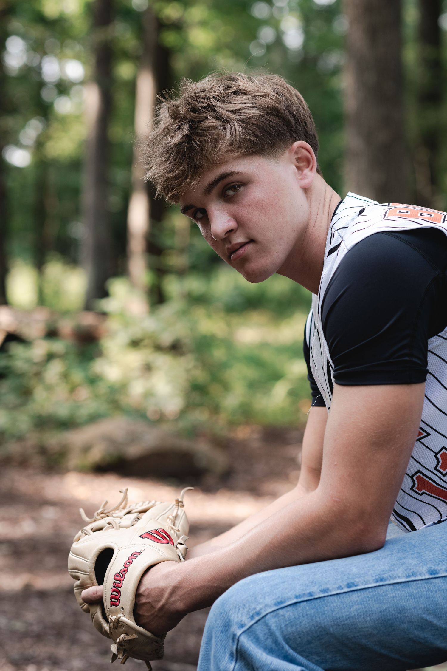 Young baseball player in jersey holds Wilson glove while sitting outdoors in wooded area.