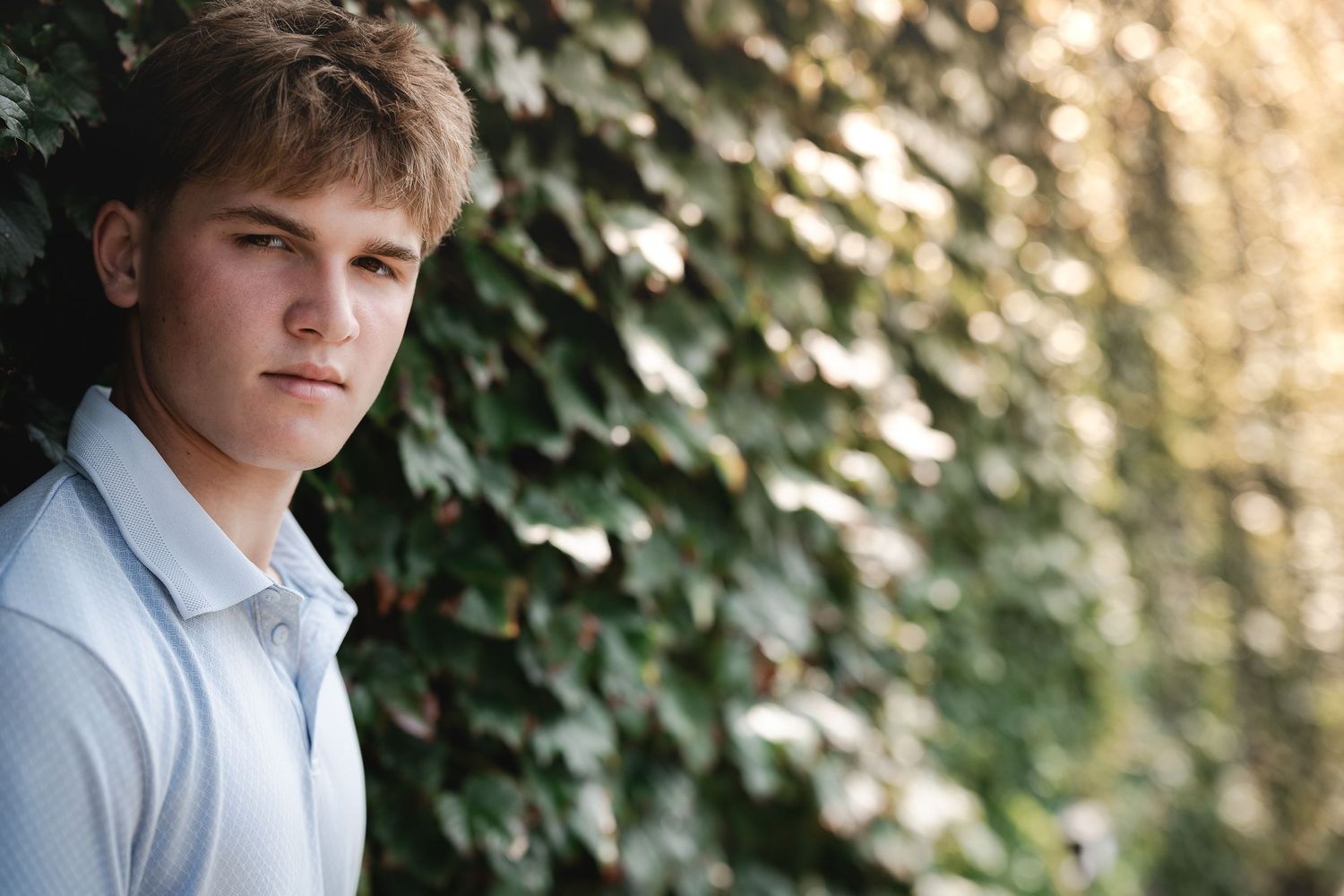 Young man in white polo shirt posing outdoors against lush green ivy wall in natural light.