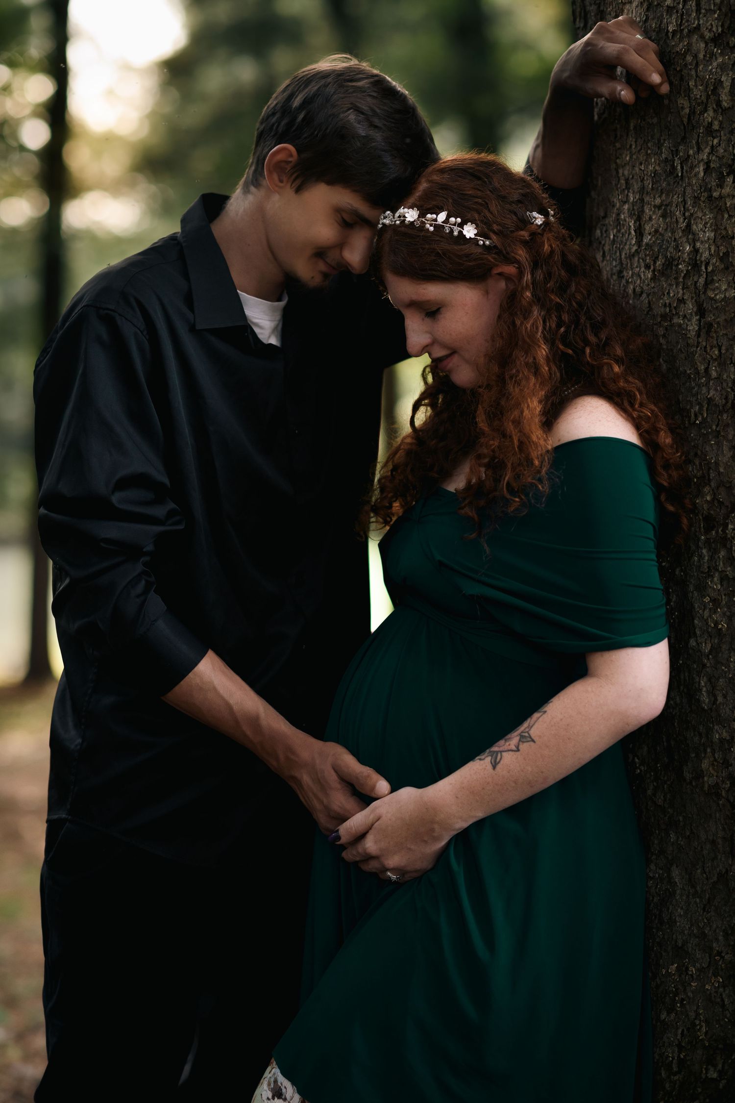 Couple sharing tender moment leaning against tree, pregnant woman in green dress with floral headband holding hands with partner.