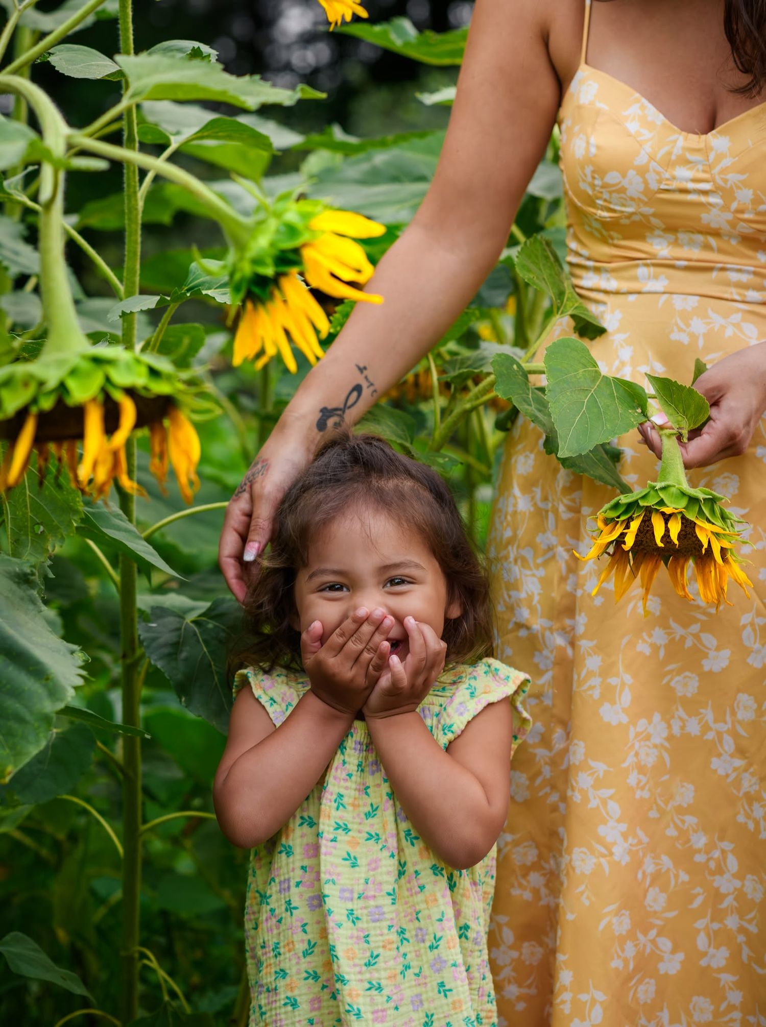 A joyful little girl covers her mouth with her hands while standing among tall sunflowers beside a woman in a yellow floral dress.