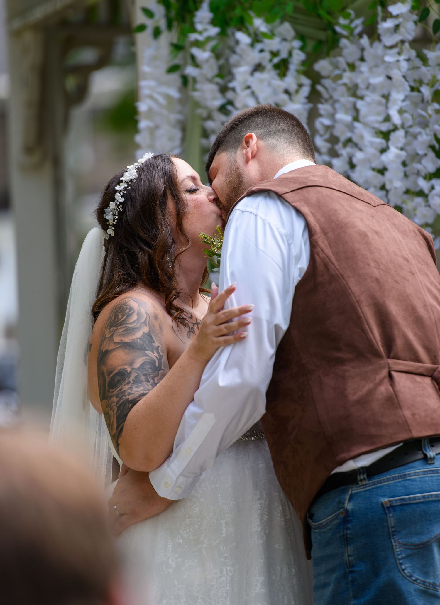 Bride and groom share their first kiss at outdoor wedding ceremony under white floral arch.