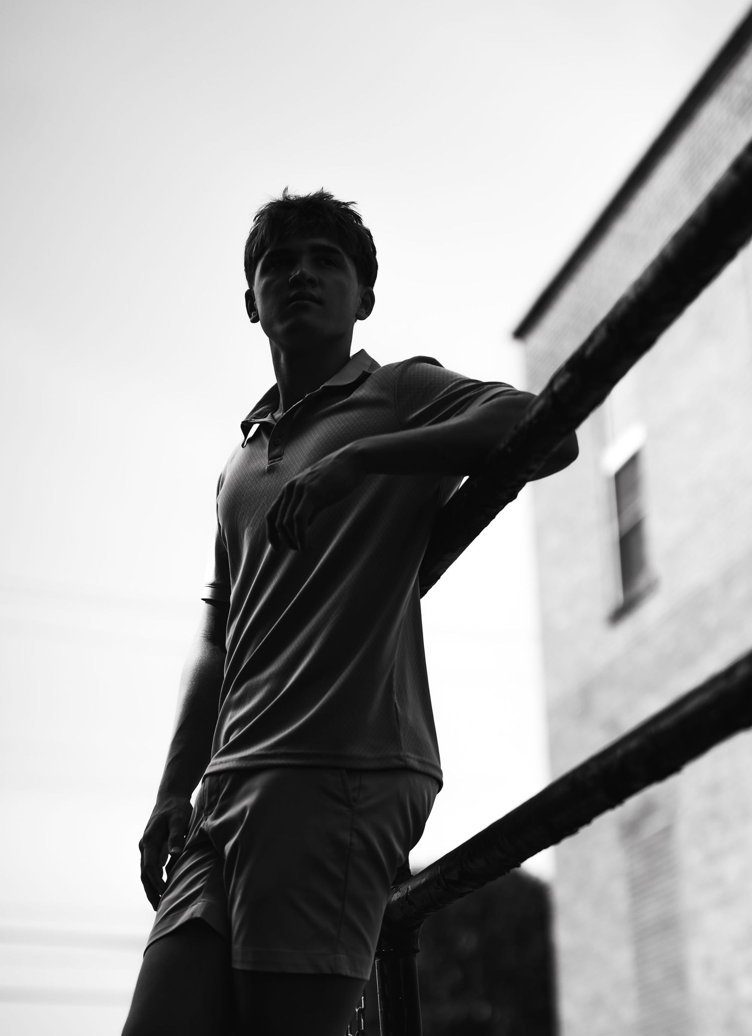 Black and white photo of a young man leaning on a railing, shot from below against a bright sky.