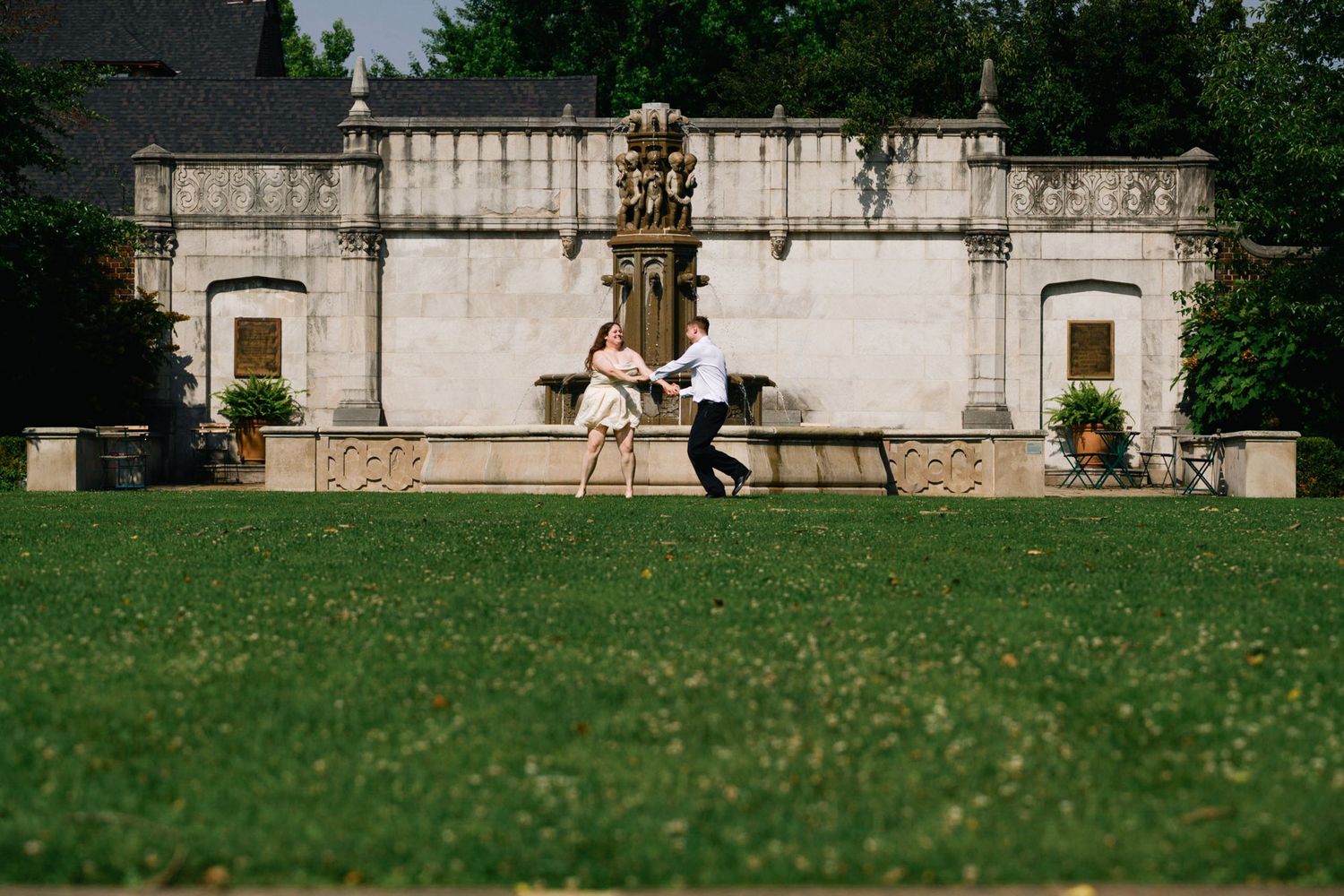 Couple dances joyfully on a green lawn in front of a grand stone fountain.