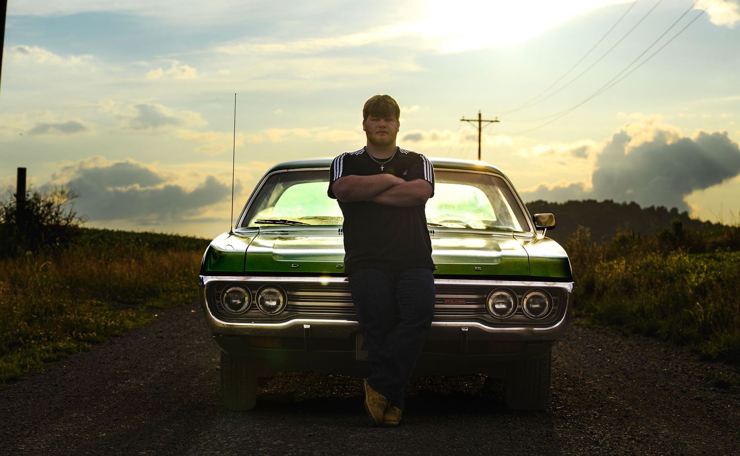 Man with crossed arms leaning against a classic green muscle car on a rural road at sunset.