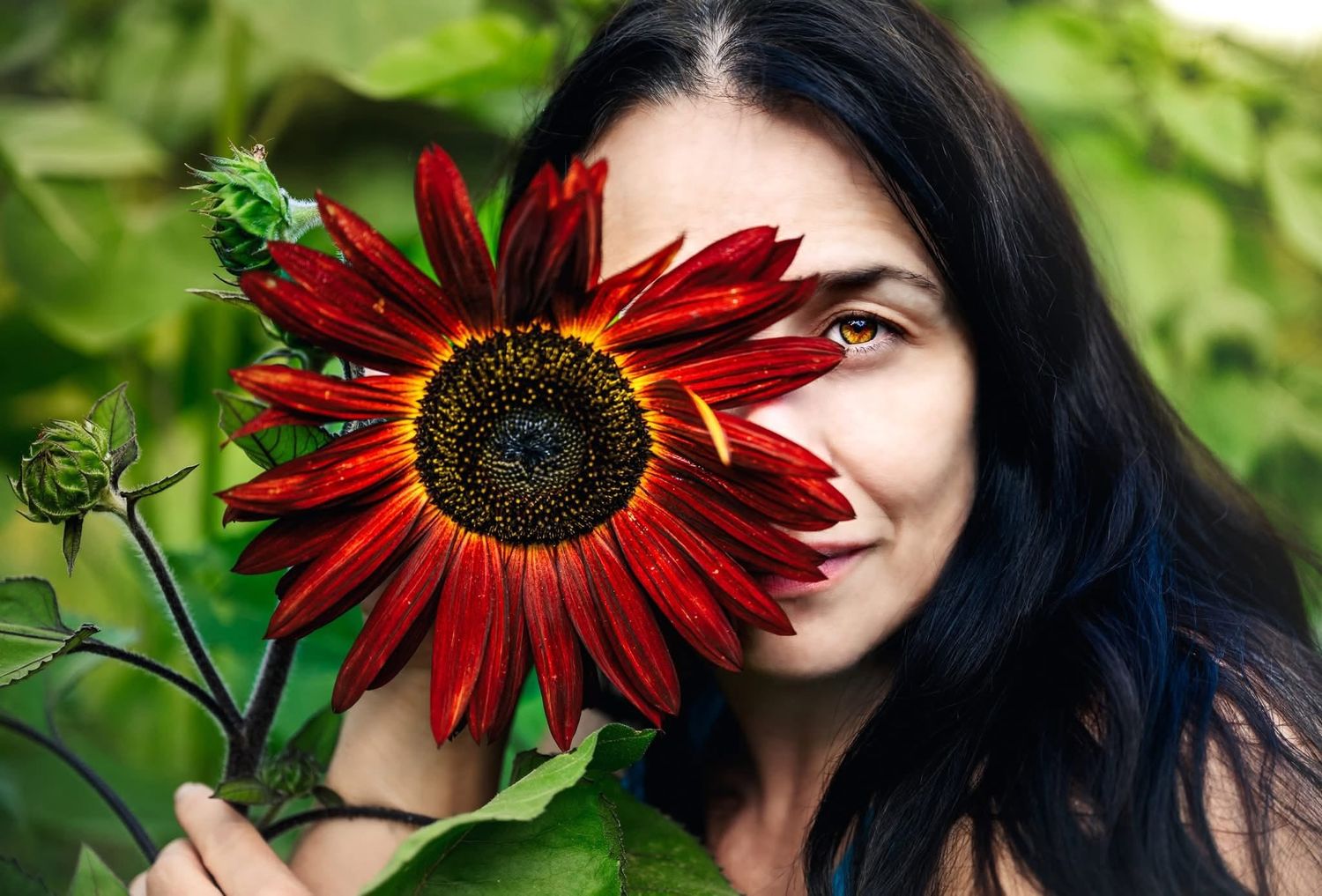 A young woman with dark hair holds a vibrant red sunflower over half her face, revealing one green eye.