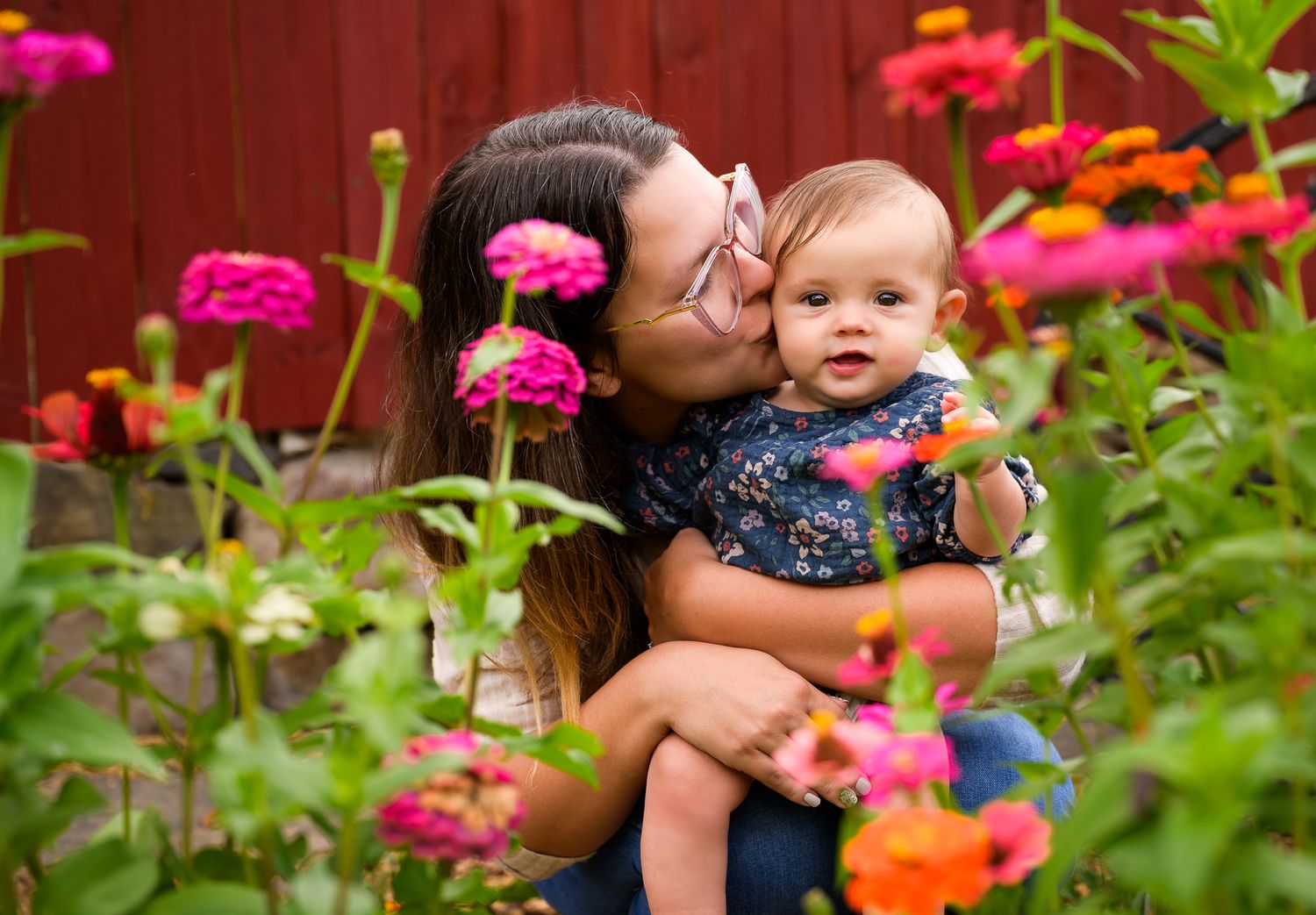 A mother kisses her baby among colorful zinnia flowers in a garden with a red barn backdrop.