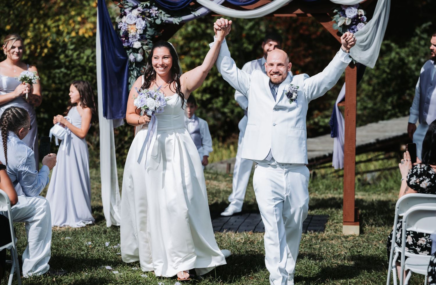 Joyful newlyweds raising hands at outdoor wedding ceremony, bride in white gown, groom in light suit.
